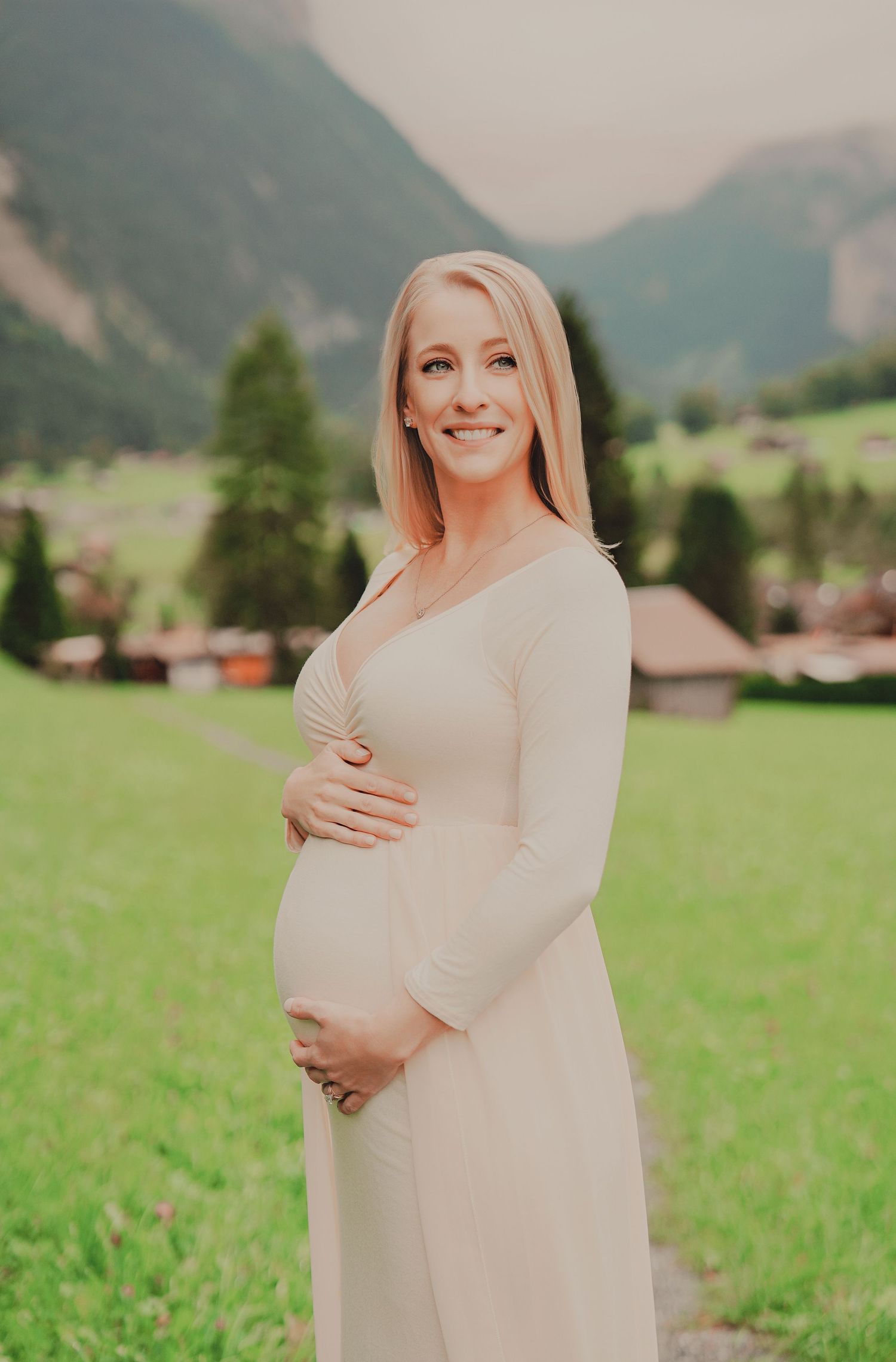 Smiling pregnant blonde woman in cream dress cradling her baby bump with lush green Swiss mountain backdrop.