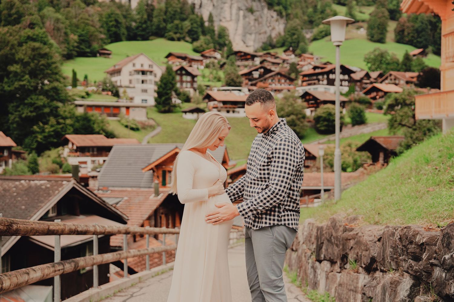 Couple lovingly looking at pregnant belly during maternity shoot with traditional Swiss chalet village backdrop.