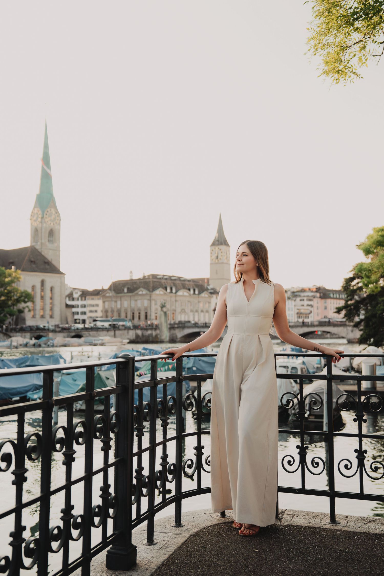 Woman in white sleeveless jumpsuit standing on ornate iron balcony overlooking Zurich river skyline.