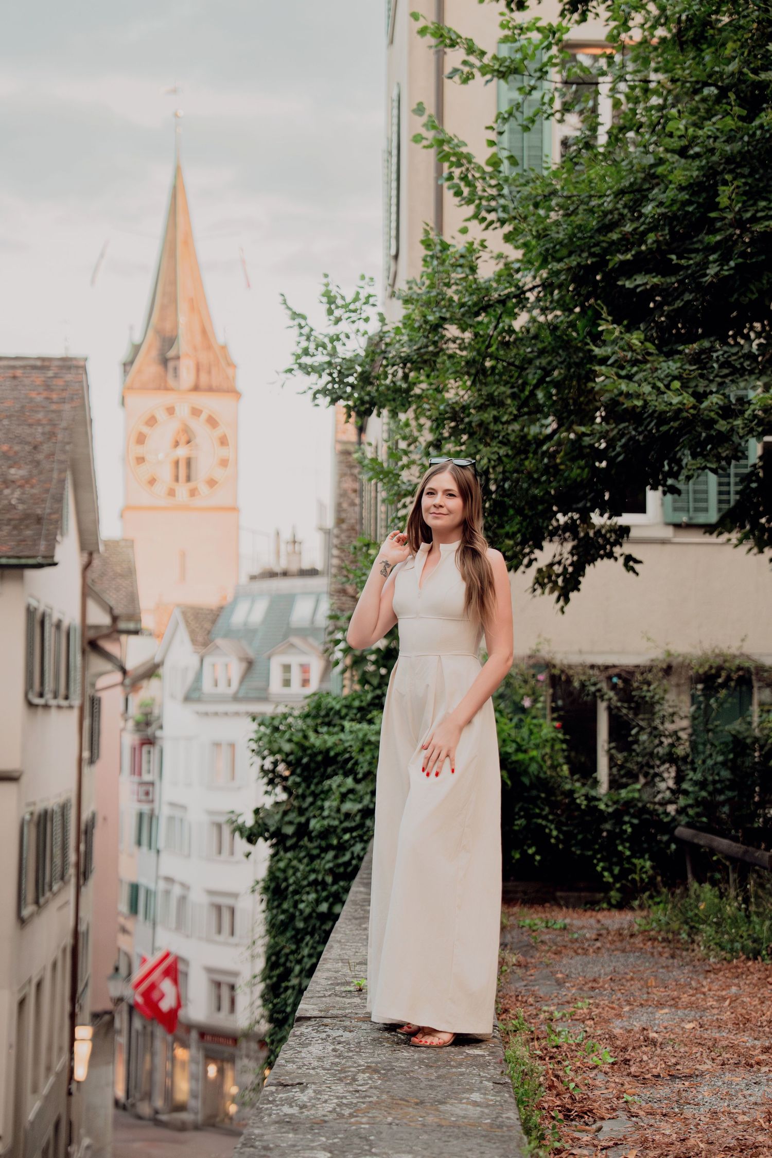 Woman in white dress stands on elevated path with St. Peter Church tower visible in Zurich.