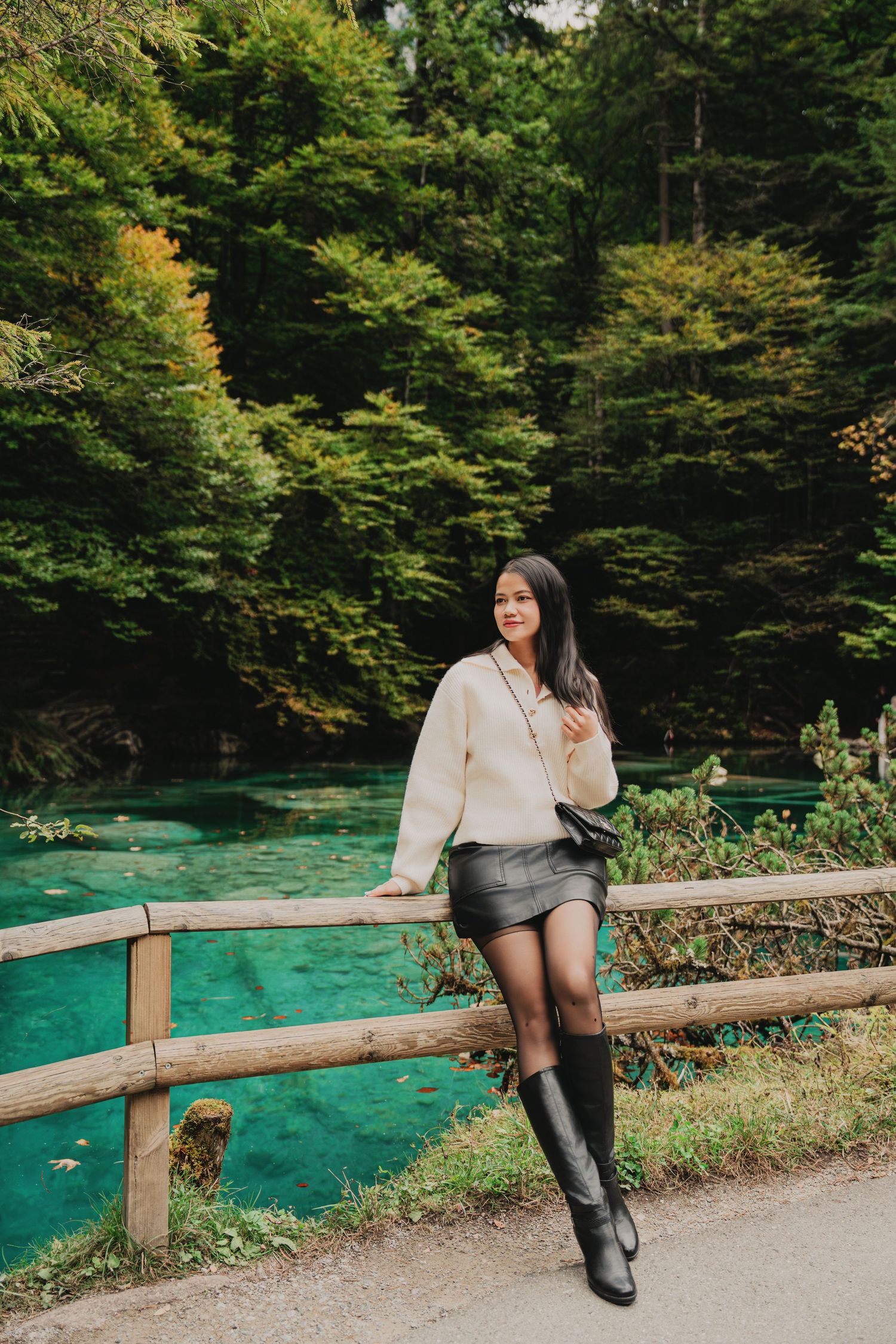 Woman in white sweater and black skirt leans on wooden fence beside stunning turquoise forest lake.