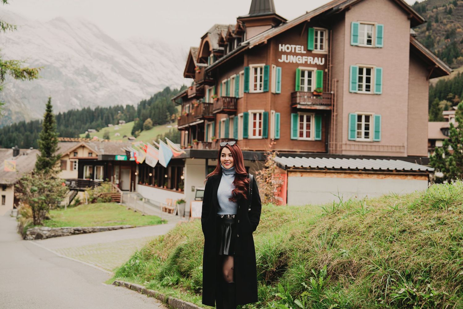 Smiling woman in black coat poses in front of Hotel Jungfrau in a scenic Swiss Alpine village.