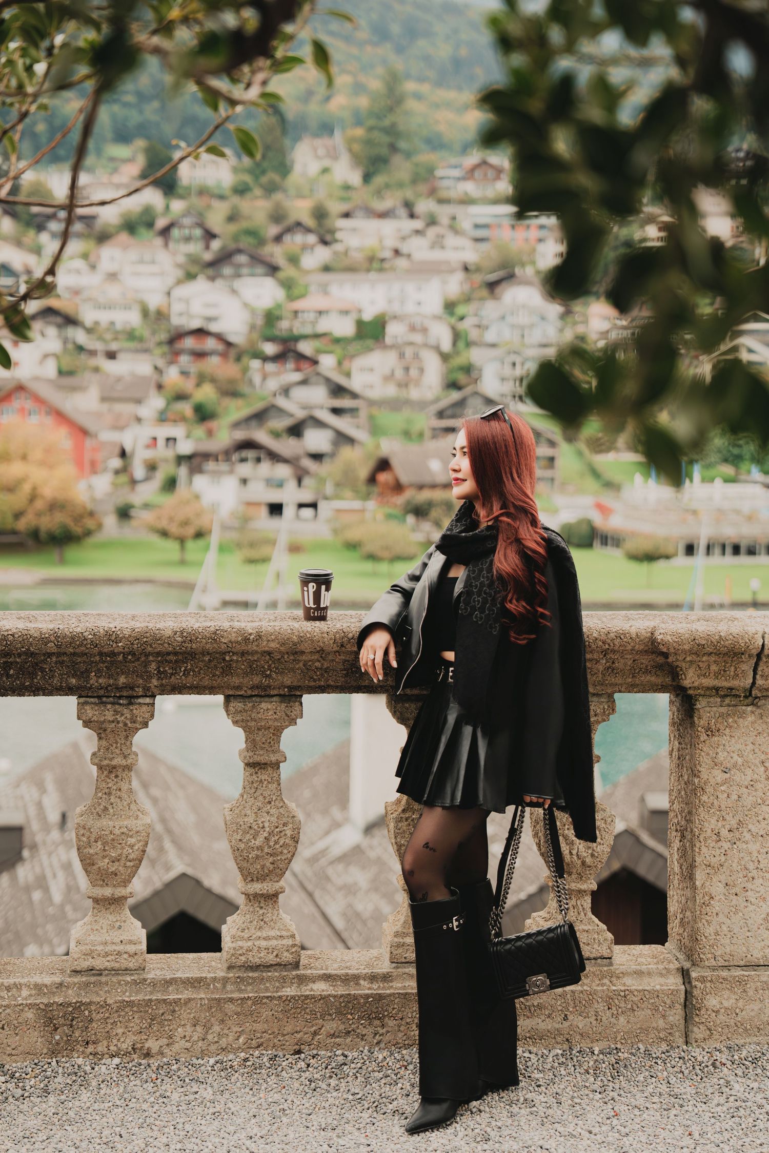 Elegant woman in black leather skirt and cape poses at stone balustrade with Swiss village backdrop.