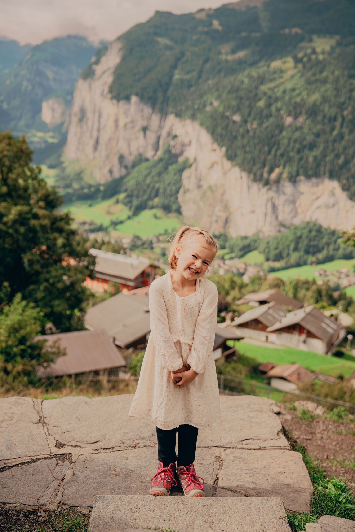 Smiling blonde girl in white dress and pink boots standing on stone wall with Alpine village below.