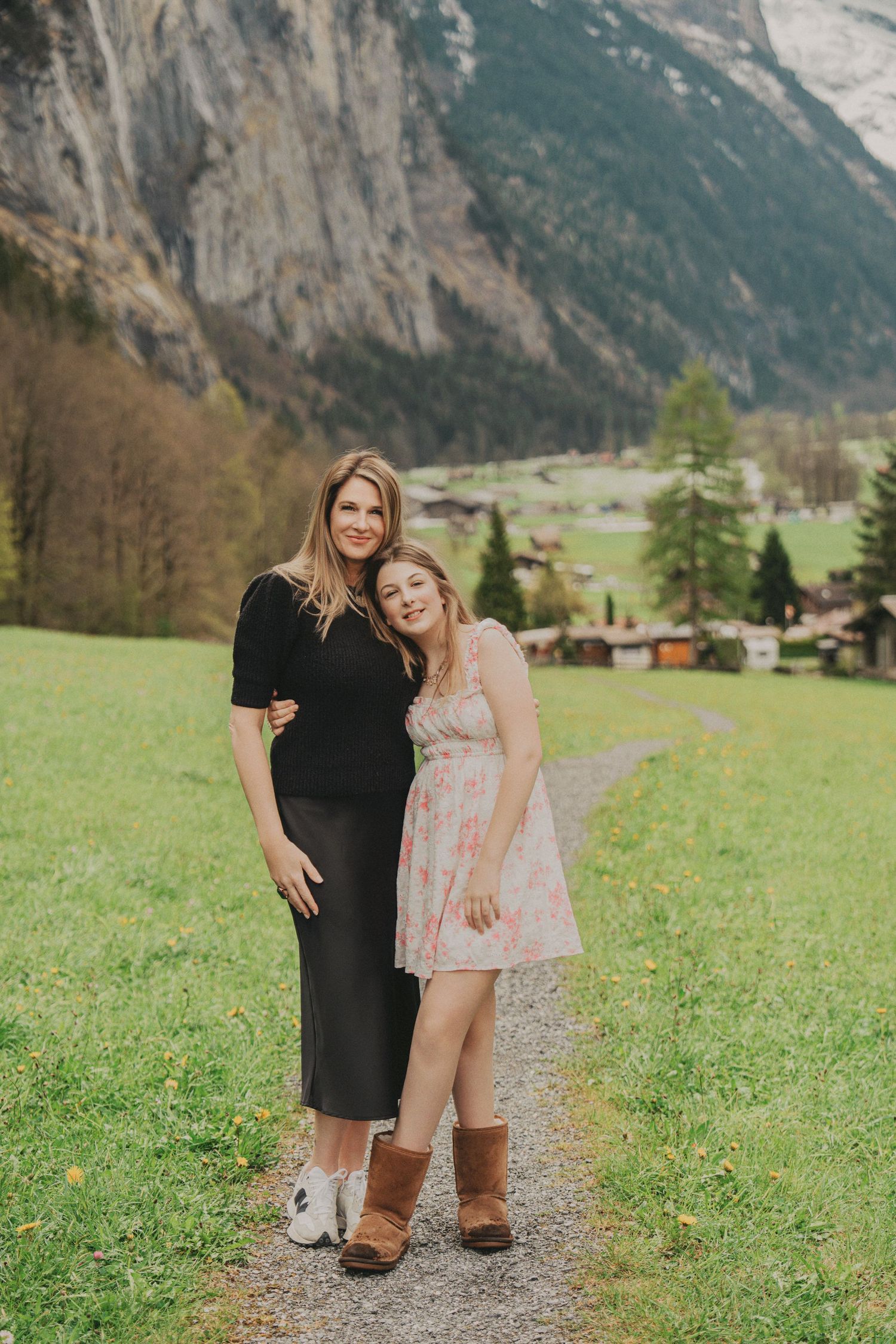 Mother and teenage daughter posing together on alpine path with Swiss mountain valley in background.