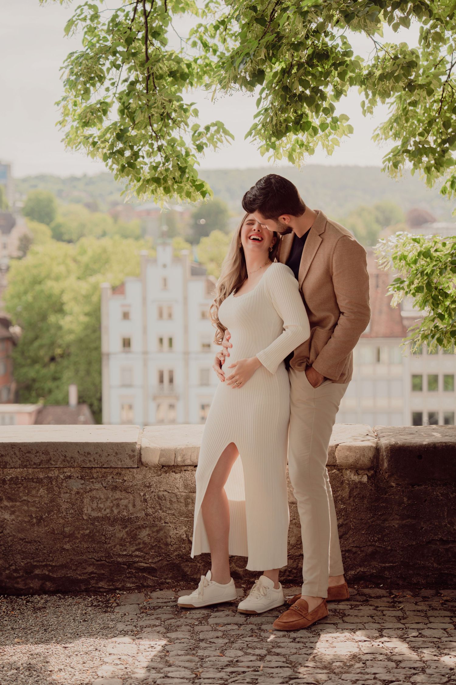 Laughing pregnant couple shares joyful moment under green tree with European city buildings behind them.
