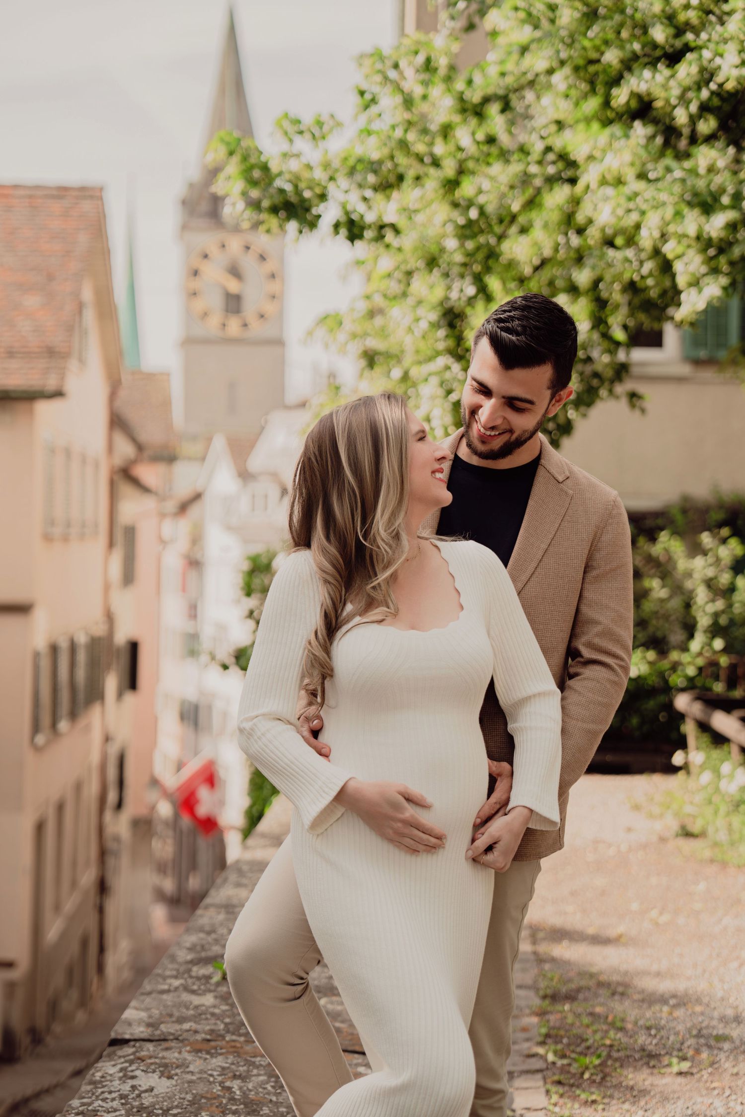 Pregnant woman in white dress gazes at partner in tan blazer during maternity shoot in Zurich old town.