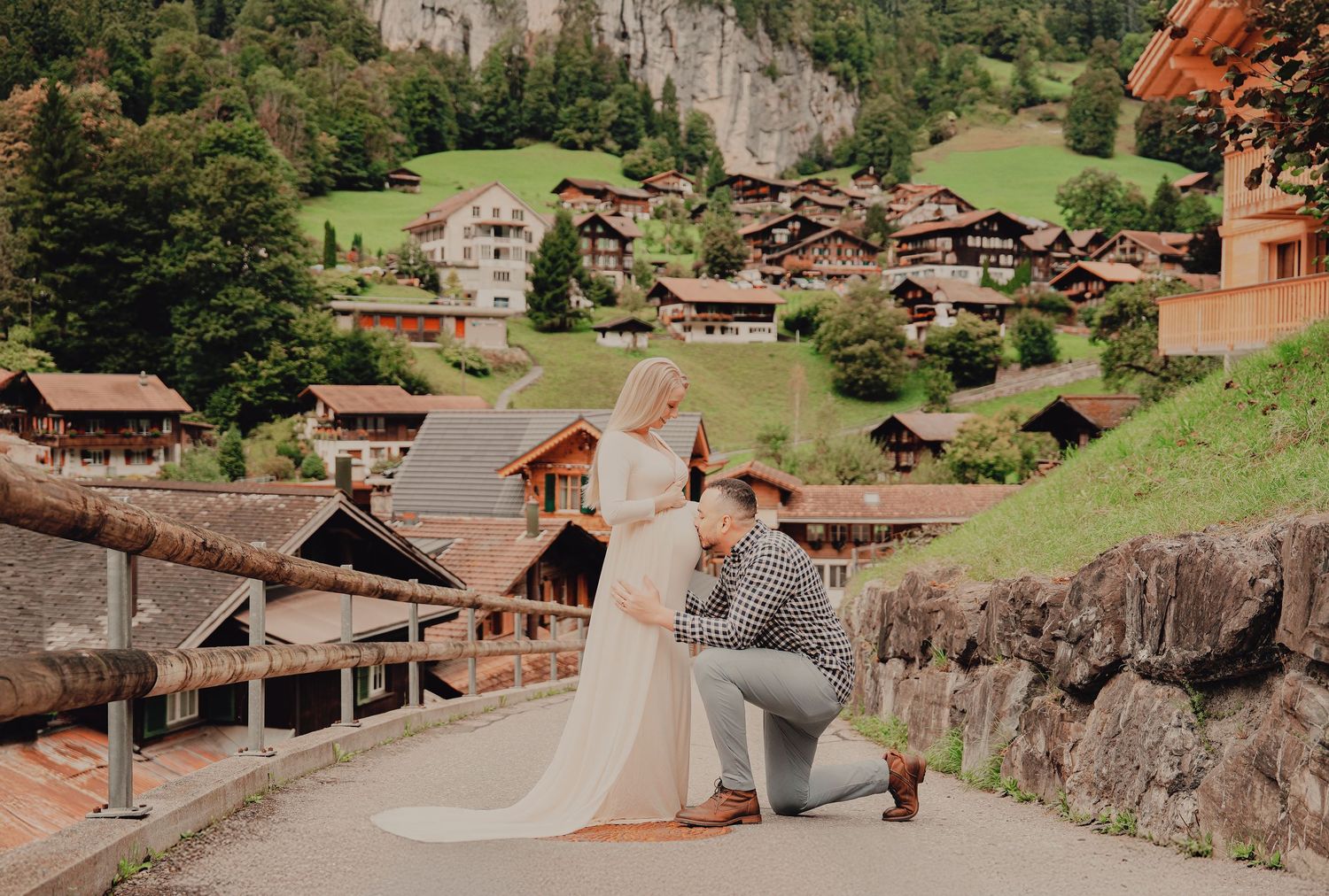 Couple poses for maternity photos in a Swiss alpine village with traditional wooden chalets in the background.