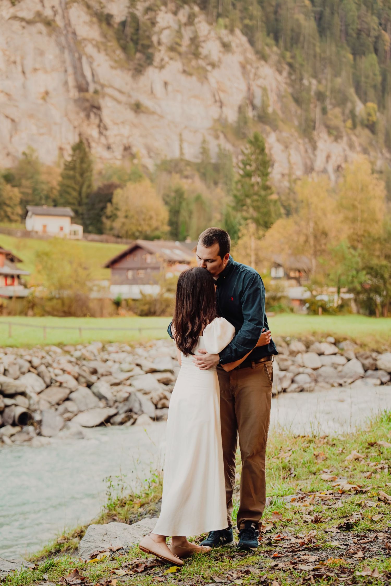 Couple sharing a tender moment by a river with Swiss alpine village and mountains.