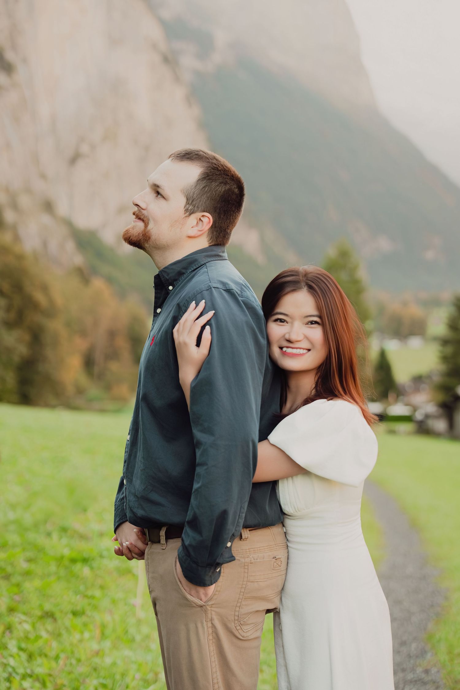 Woman in white dress hugging man from behind while smiling in alpine meadow.