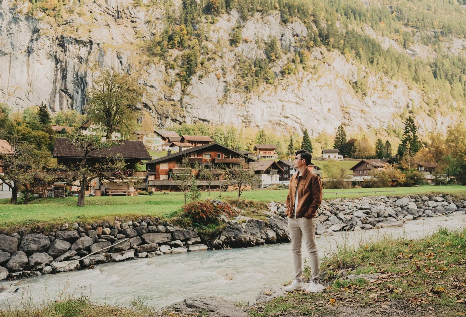 Man posing beside a glacial river with charming Swiss village chalets and autumn mountain scenery.