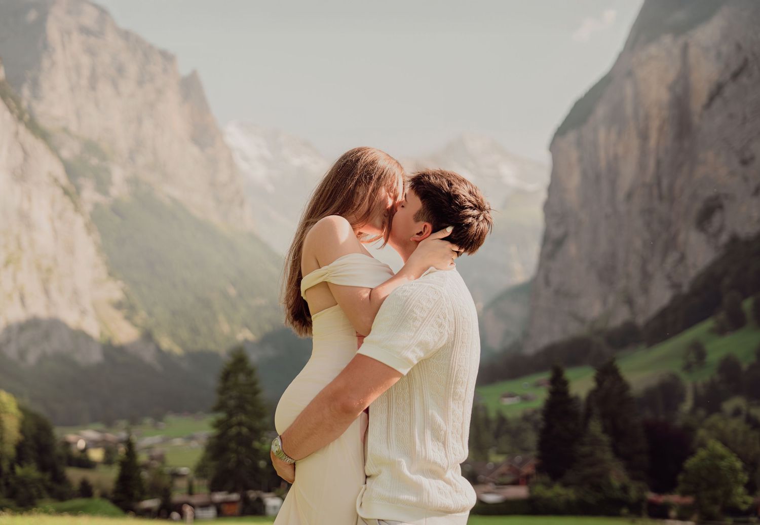Couple kissing as man lifts woman in white off-shoulder dress against stunning Swiss mountain scenery.