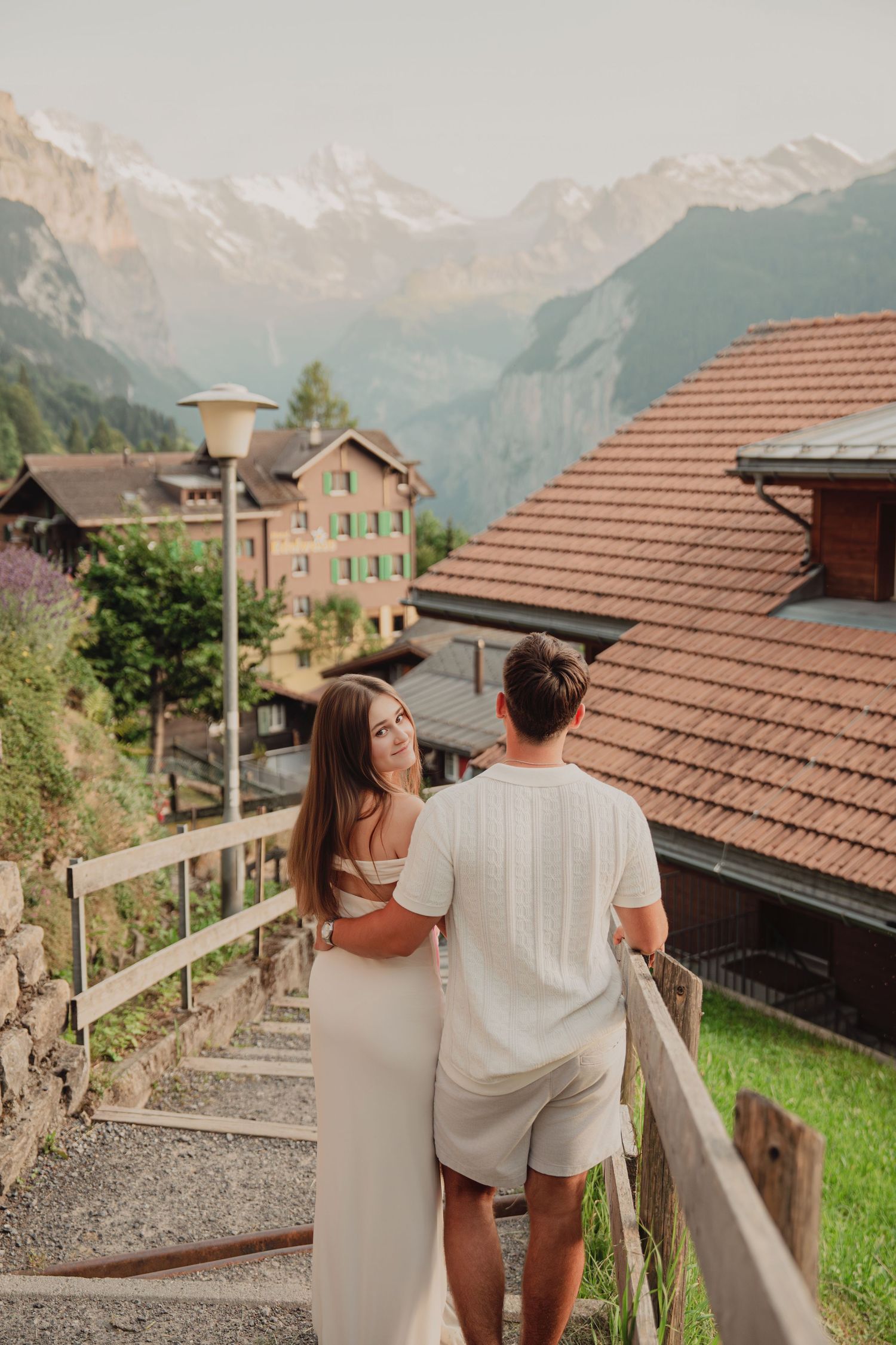 Woman in white dress embracing man on wooden staircase in idyllic Swiss mountain village at sunset.