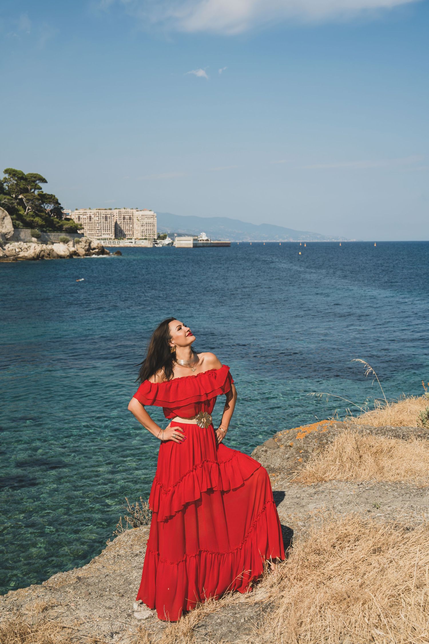 Woman in flowing red off-shoulder tiered dress standing on rocky Mediterranean coastline under blue sky.