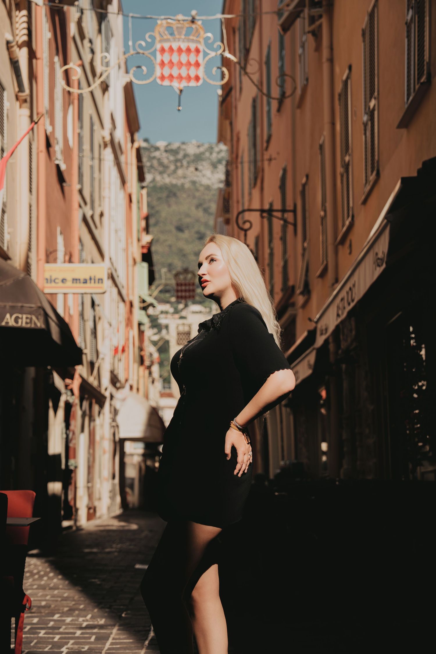 Blonde woman in black dress posing in narrow Monaco street with colorful buildings.