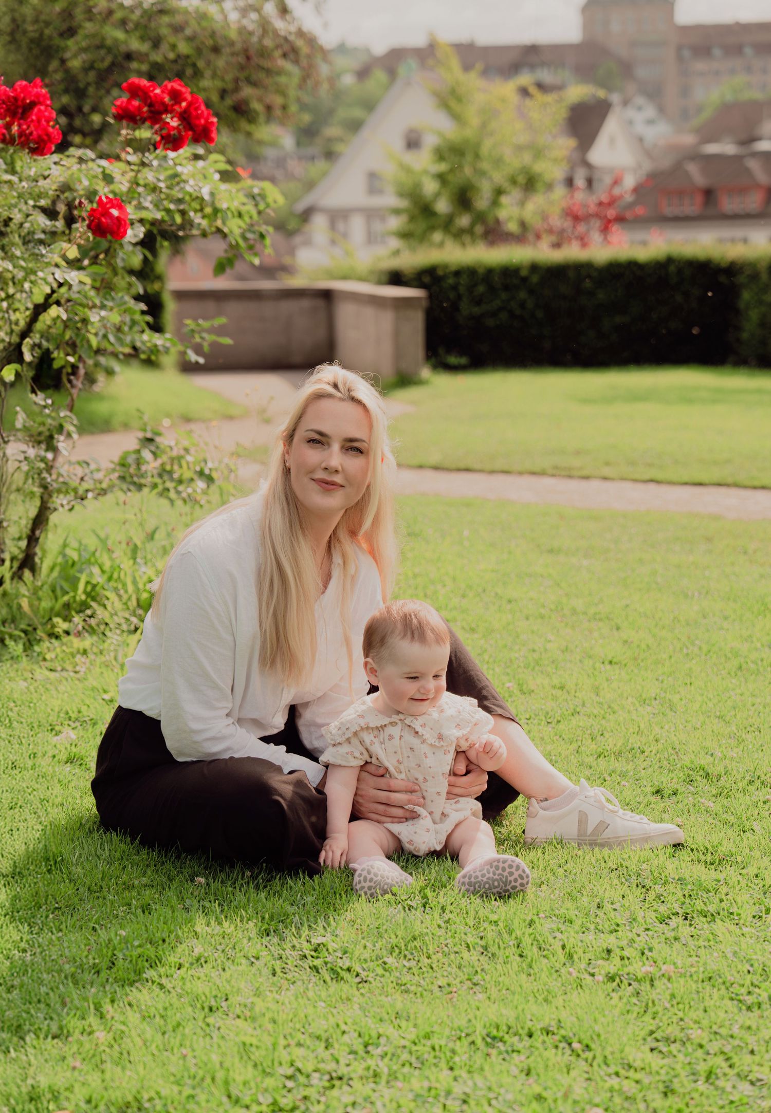 Blonde mother sitting on grass with baby near blooming red roses in European garden setting.