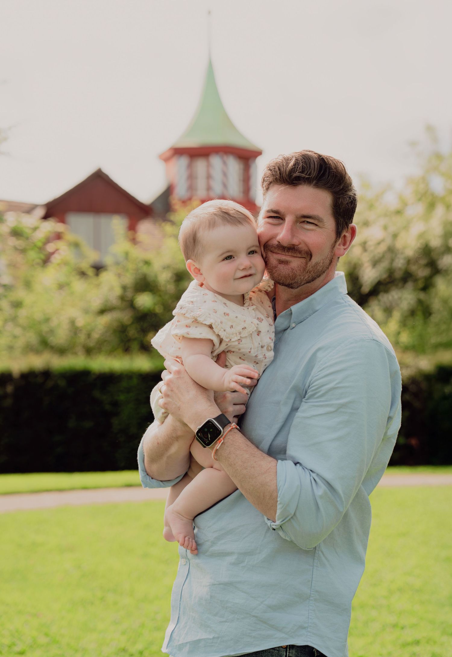 Smiling man in light blue shirt holding a happy baby outdoors in a green park with a red building behind.