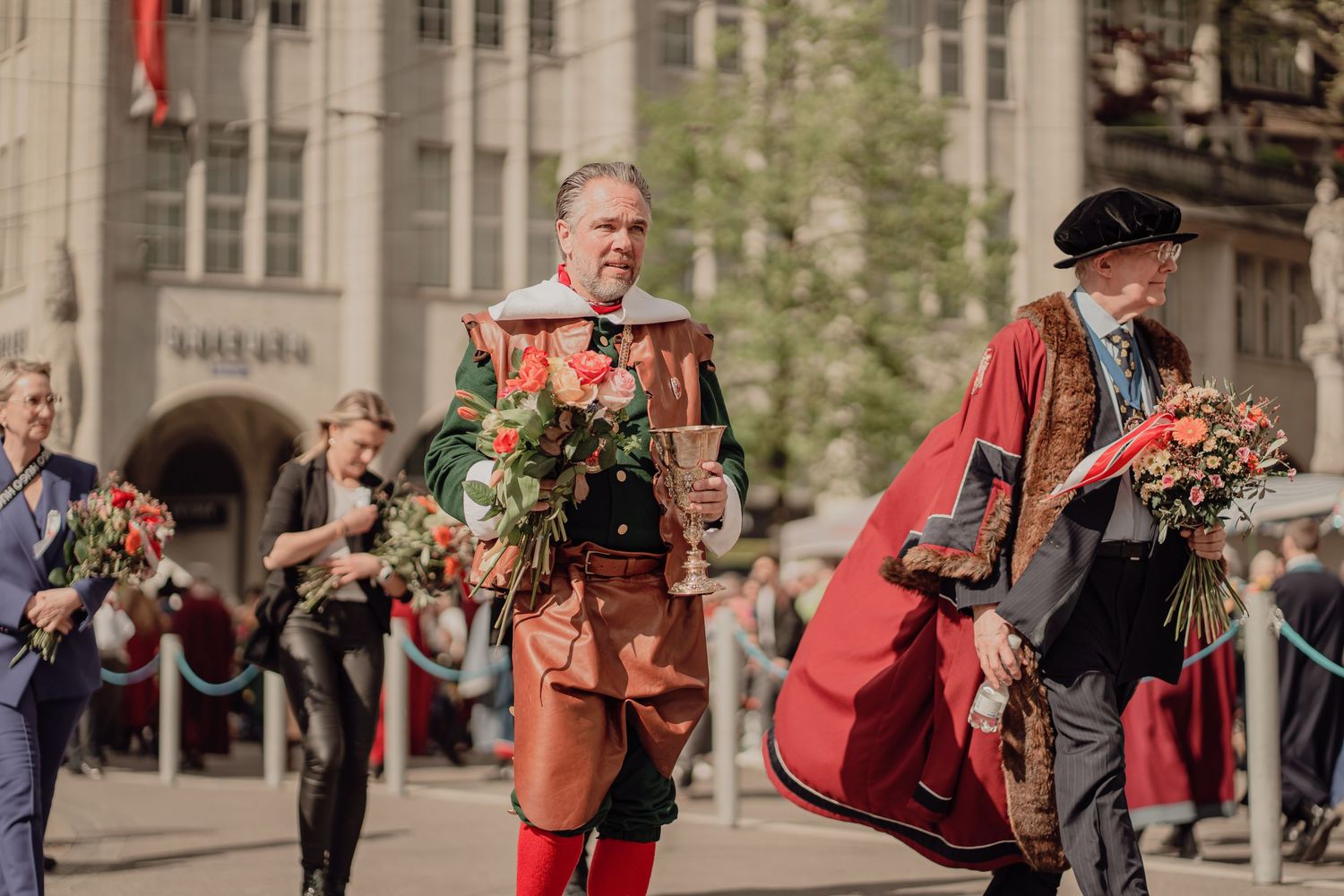 Two men in elaborate historical costumes carry flower bouquets during a traditional European parade.