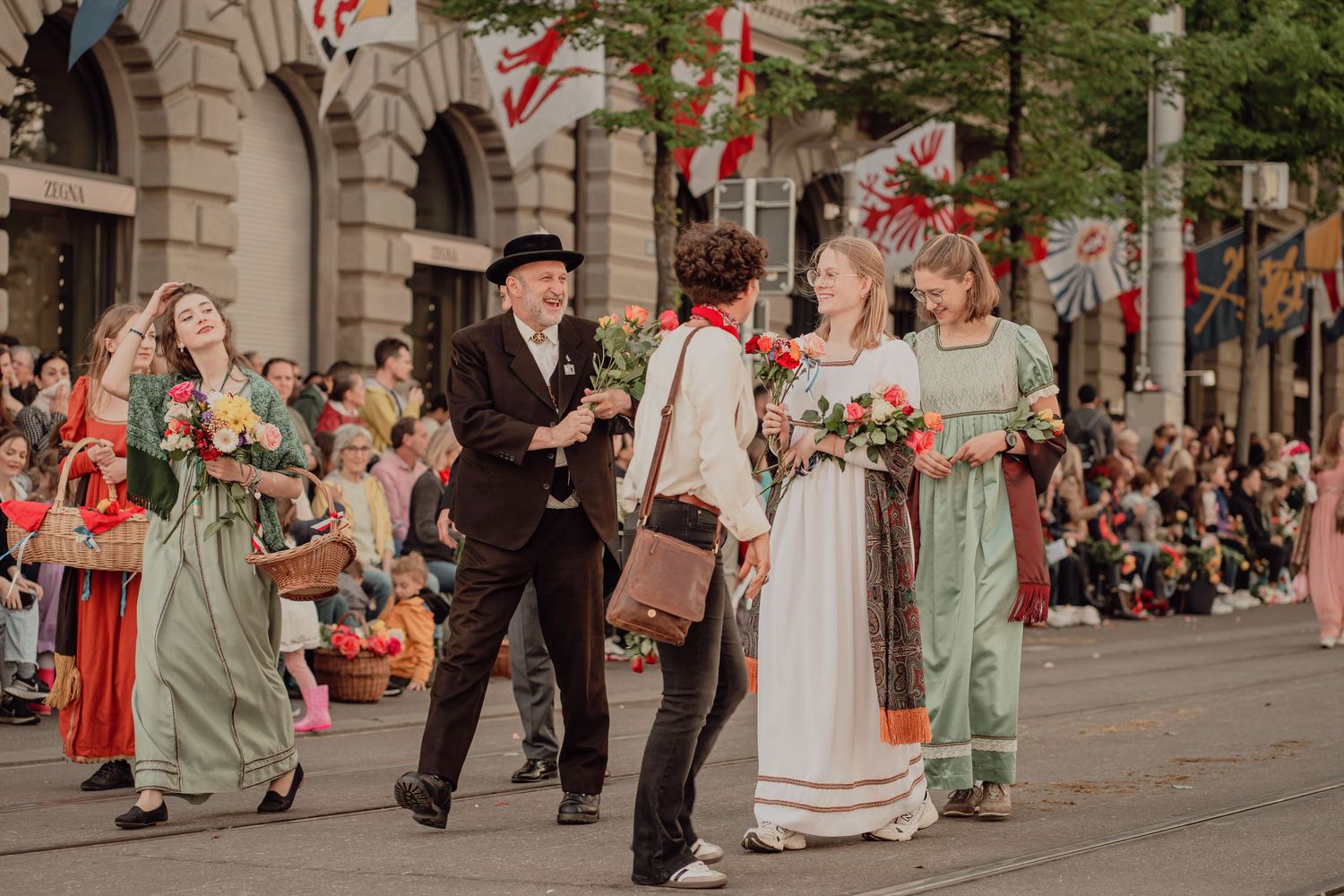 Performers in traditional folk costumes interact and carry flowers during a colorful street parade.