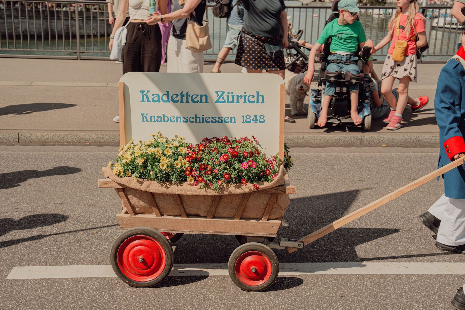 A decorated wagon with red flowers and a sign reading Kadetten Zürich Knabenschiessen 1348 at a parade.