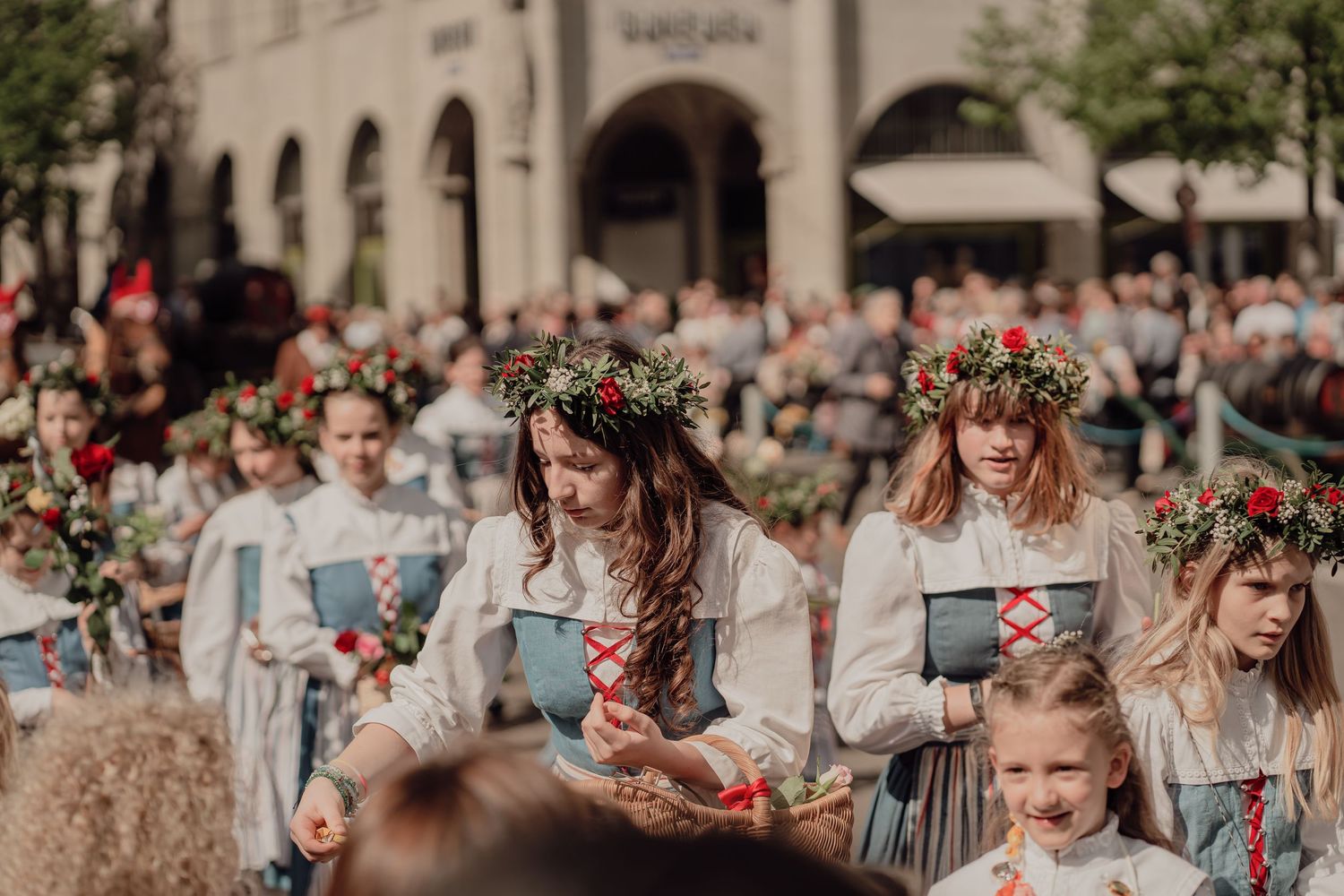 Girls wearing red floral wreaths and white folk costumes carry books in traditional cultural procession.