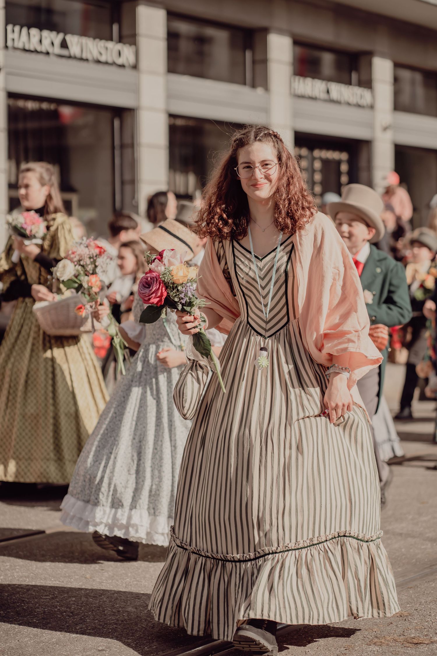 Young woman in a striped historical dress carries roses past a Harry Winston store in a parade.