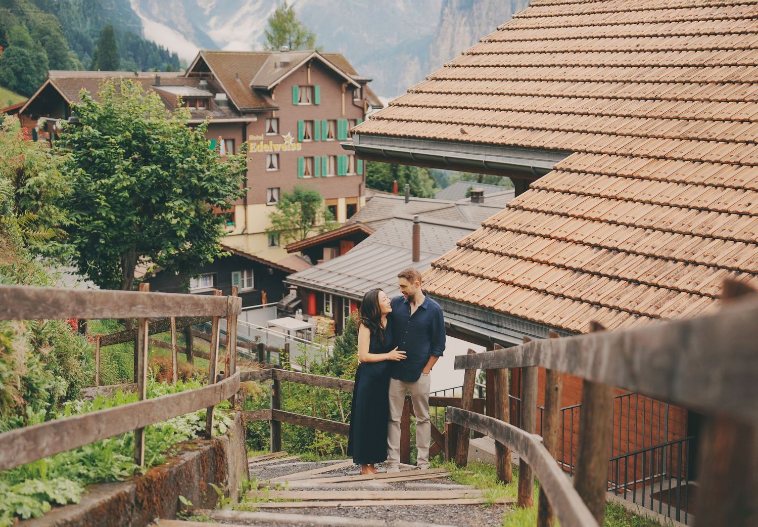A couple shares a tender moment on rustic wooden steps in a scenic Swiss alpine village setting.