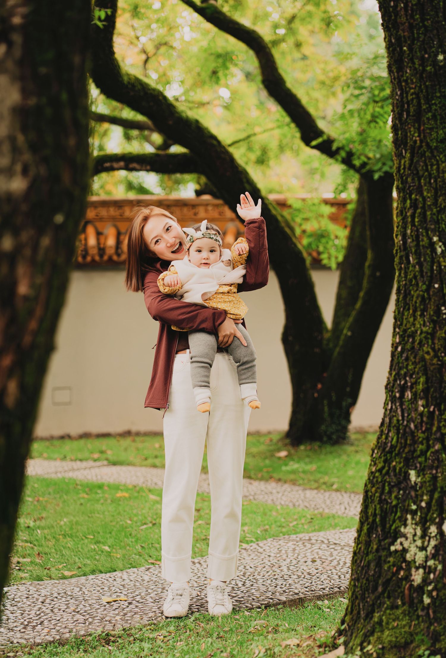 Young mother lifting a happy baby girl outdoors among green trees in a park.