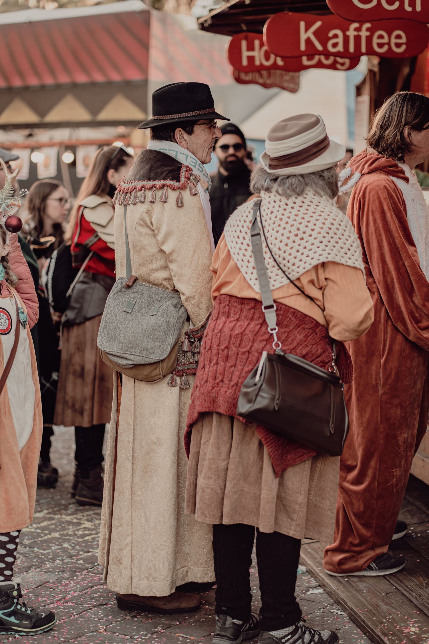 People in historical costumes at outdoor carnival market with Kaffee sign.