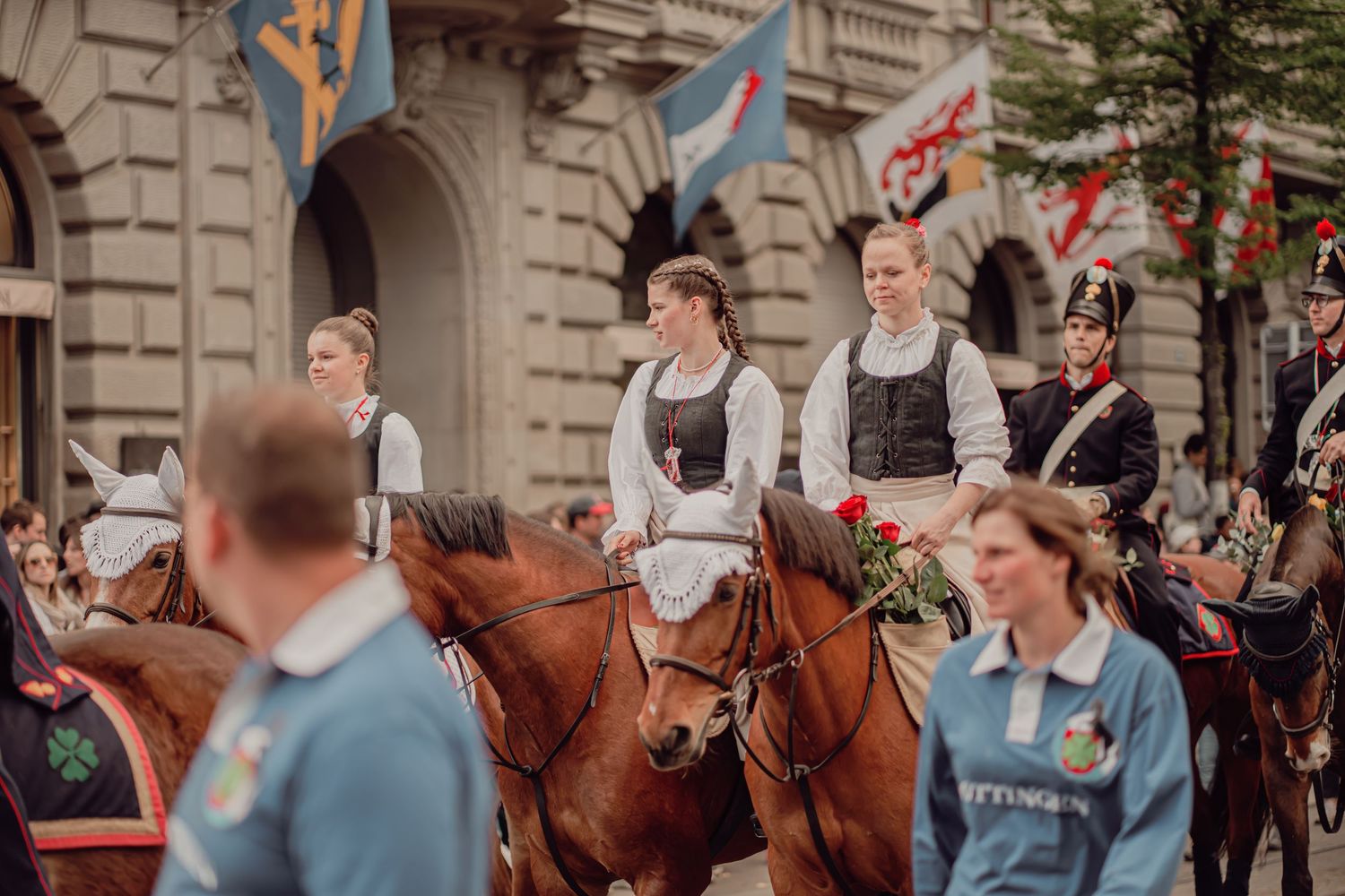 Young women in traditional white folk costumes ride horses carrying roses in a cultural parade.