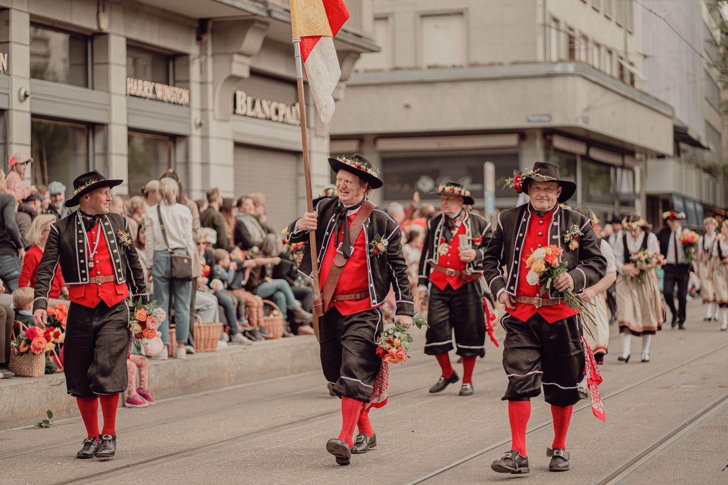 Men in traditional Swiss folk costumes with red and black attire march in a festive parade.