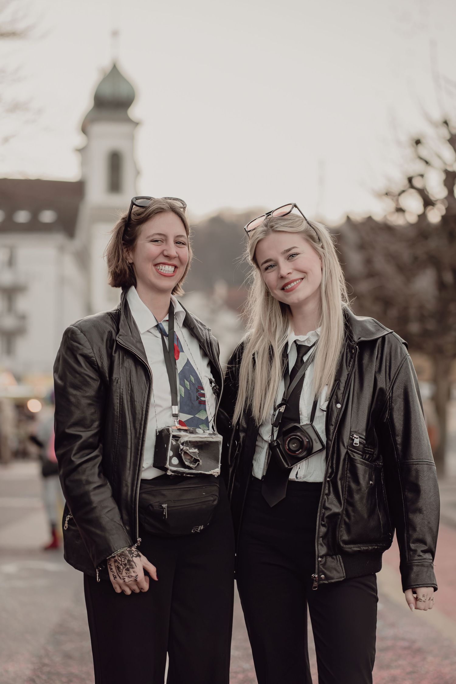 Two smiling women in matching black leather jackets and ties carry film cameras at a street carnival event at dusk.