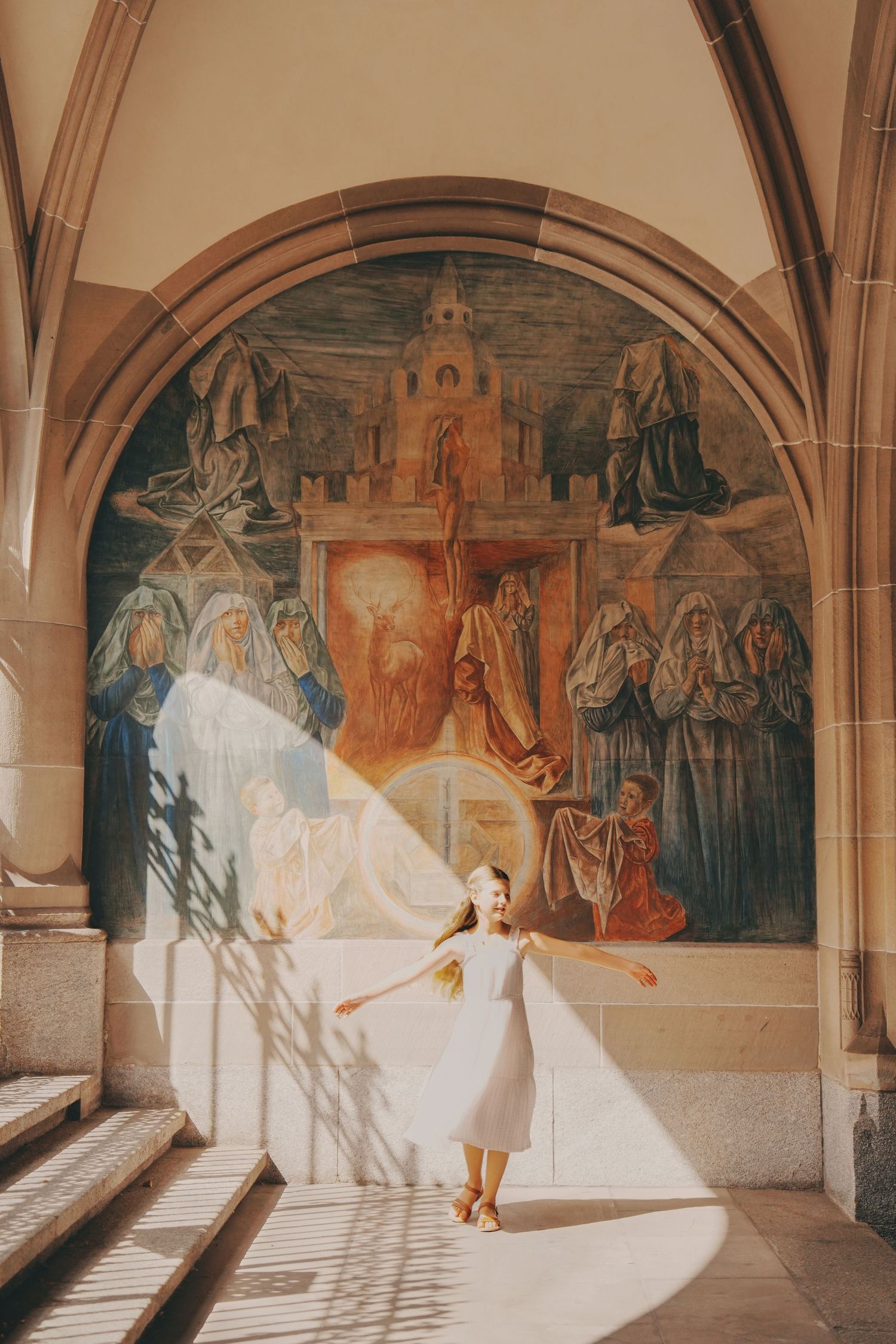 Young girl in white dress twirling before grand religious fresco in arched church corridor.