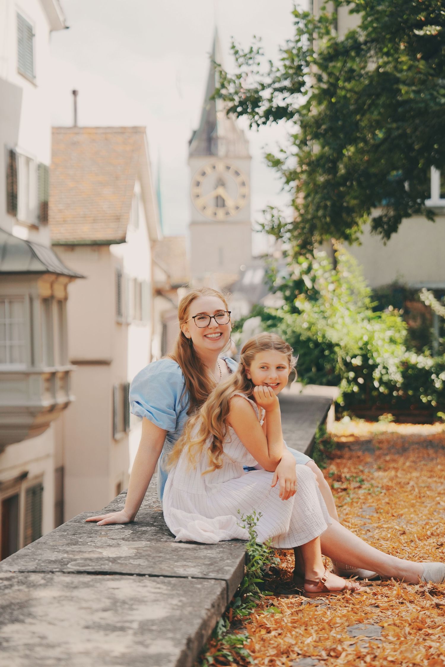 A woman wearing glasses and a girl in a white dress sit together on a stone wall with a church tower behind.