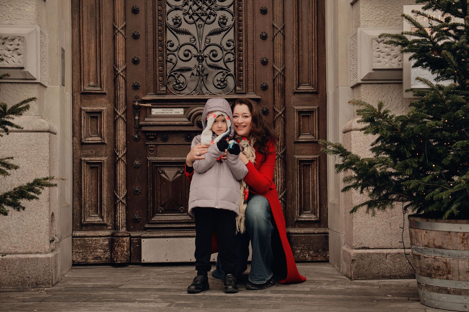 Mother in red coat crouching with young child in front of decorative vintage wooden door.