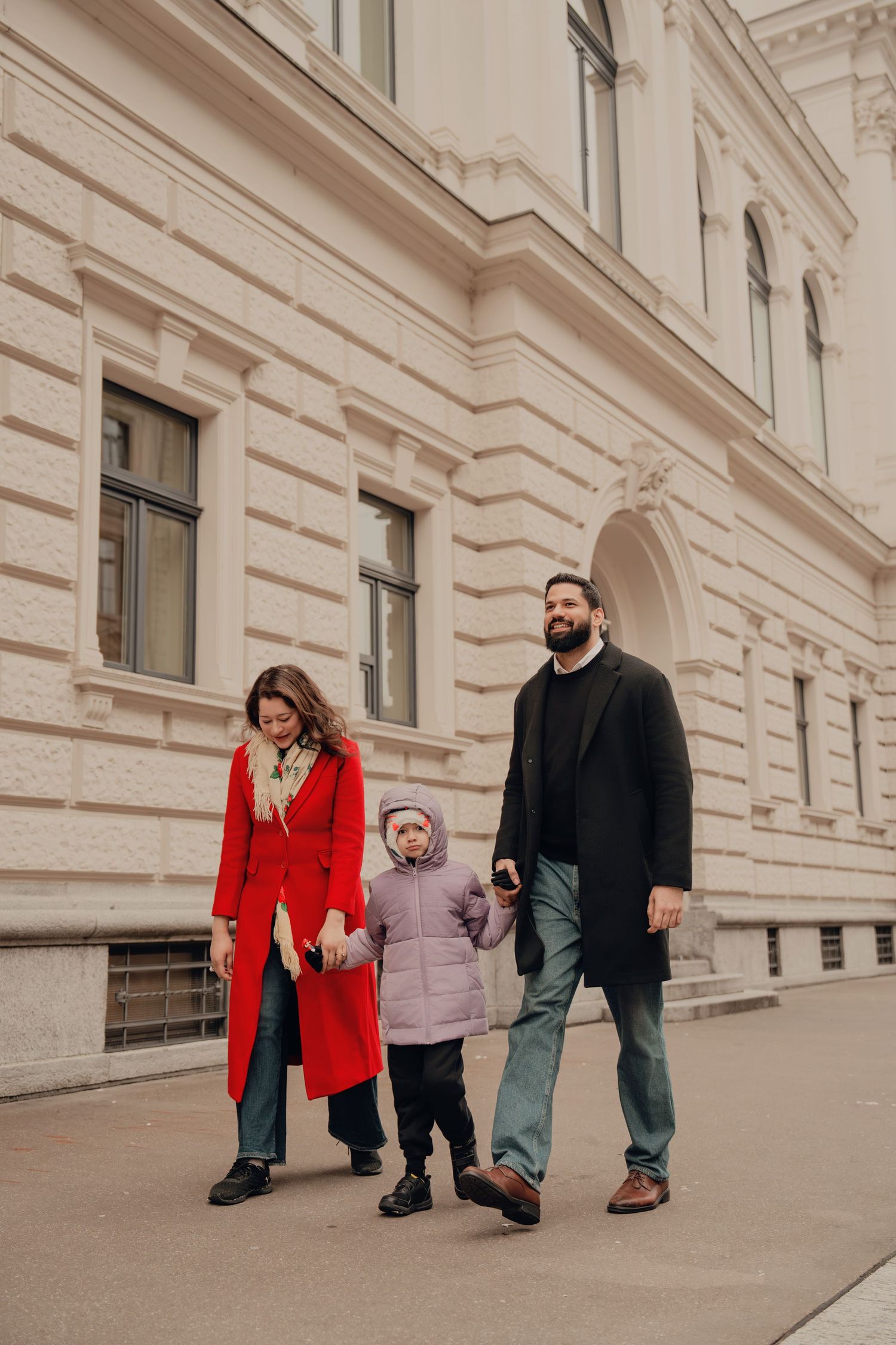 Family of three strolling together along a historic neoclassical building in winter coats.
