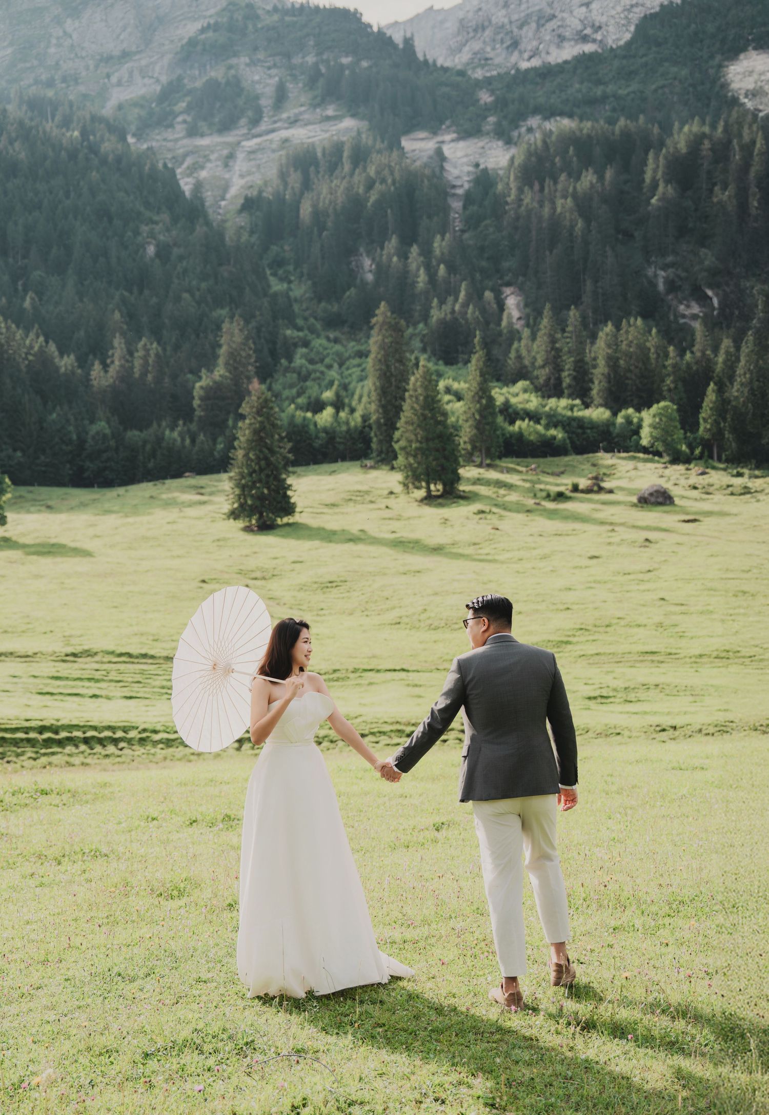 Newlyweds walk hand in hand across green alpine meadow, bride holding white parasol with forested mountains behind.