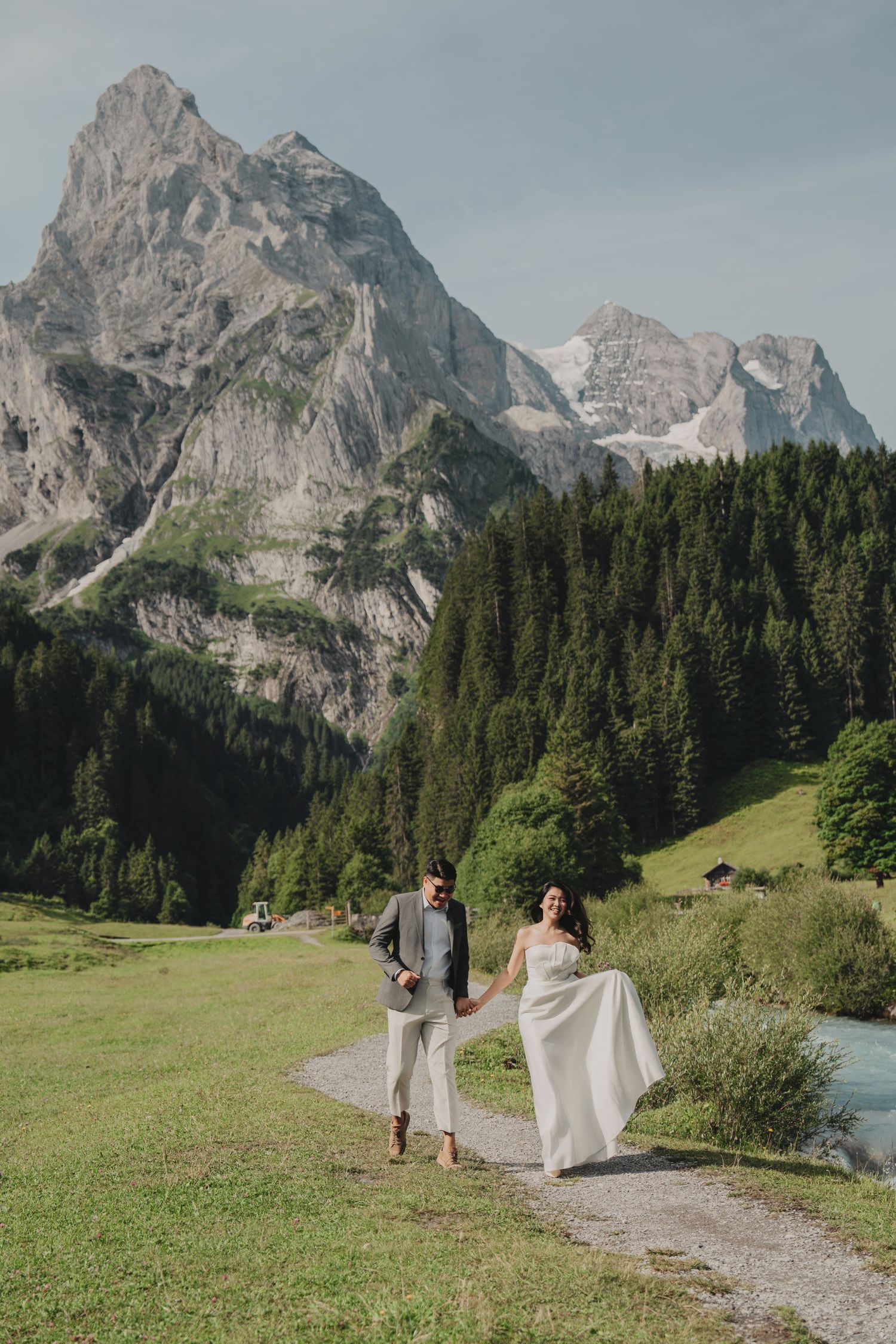 Bride's gown flowing in the breeze as wedding couple walks hand in hand along an alpine path with mountain views.