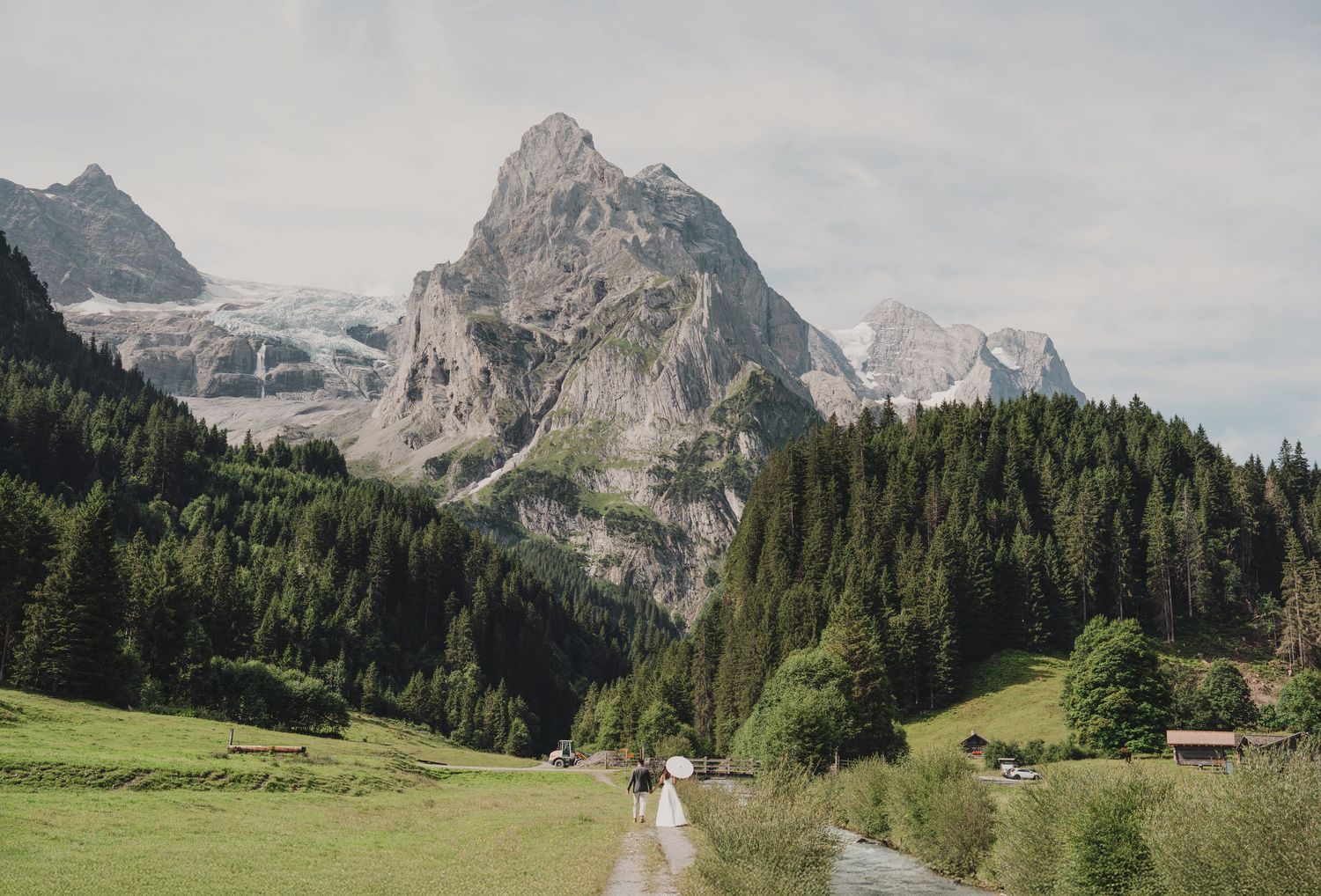 Couple in wedding attire walking along a mountain stream path with snow-capped Alpine peaks in the background.
