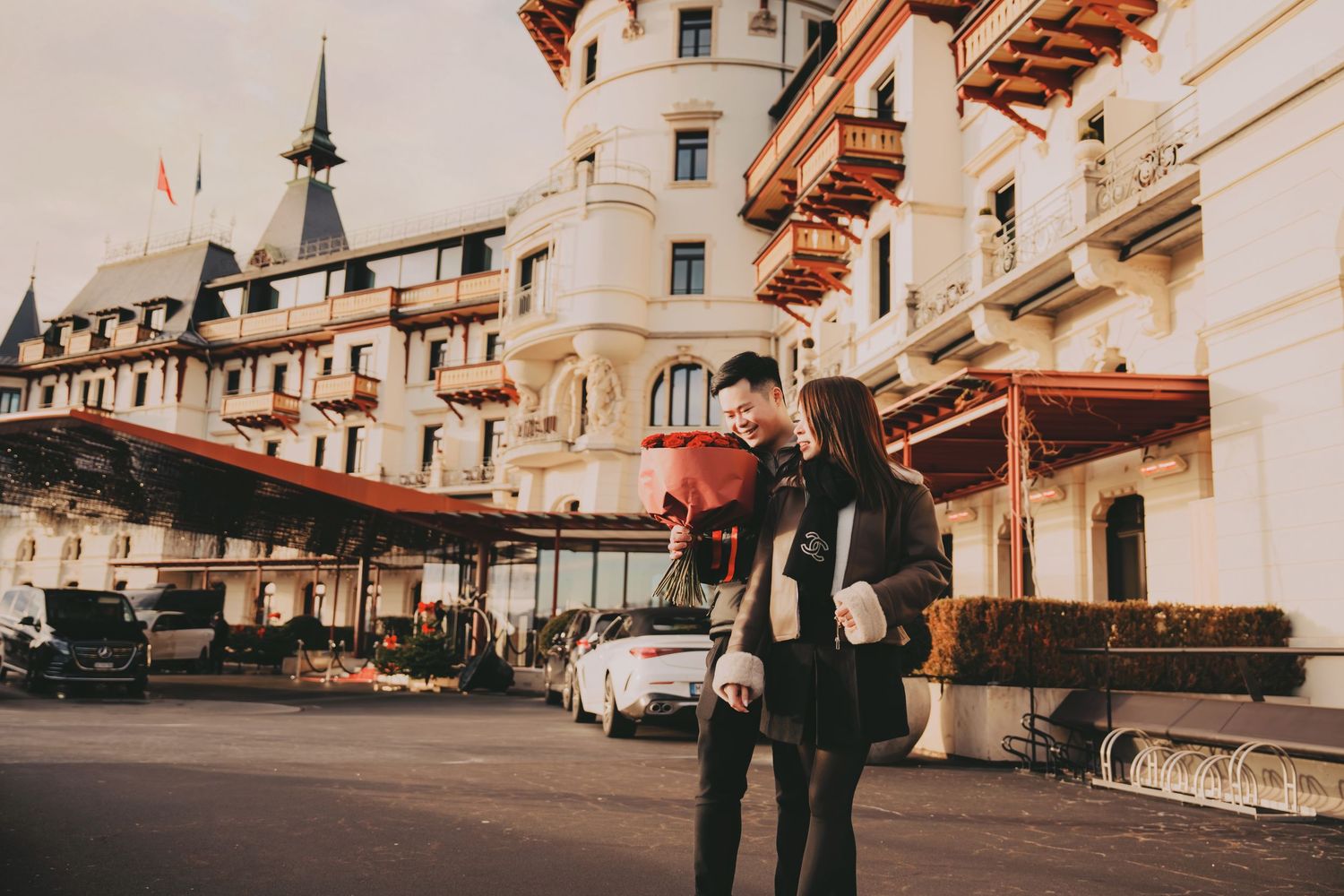 Couple embracing outside a grand European castle-style hotel with red balloons on a cloudy day.