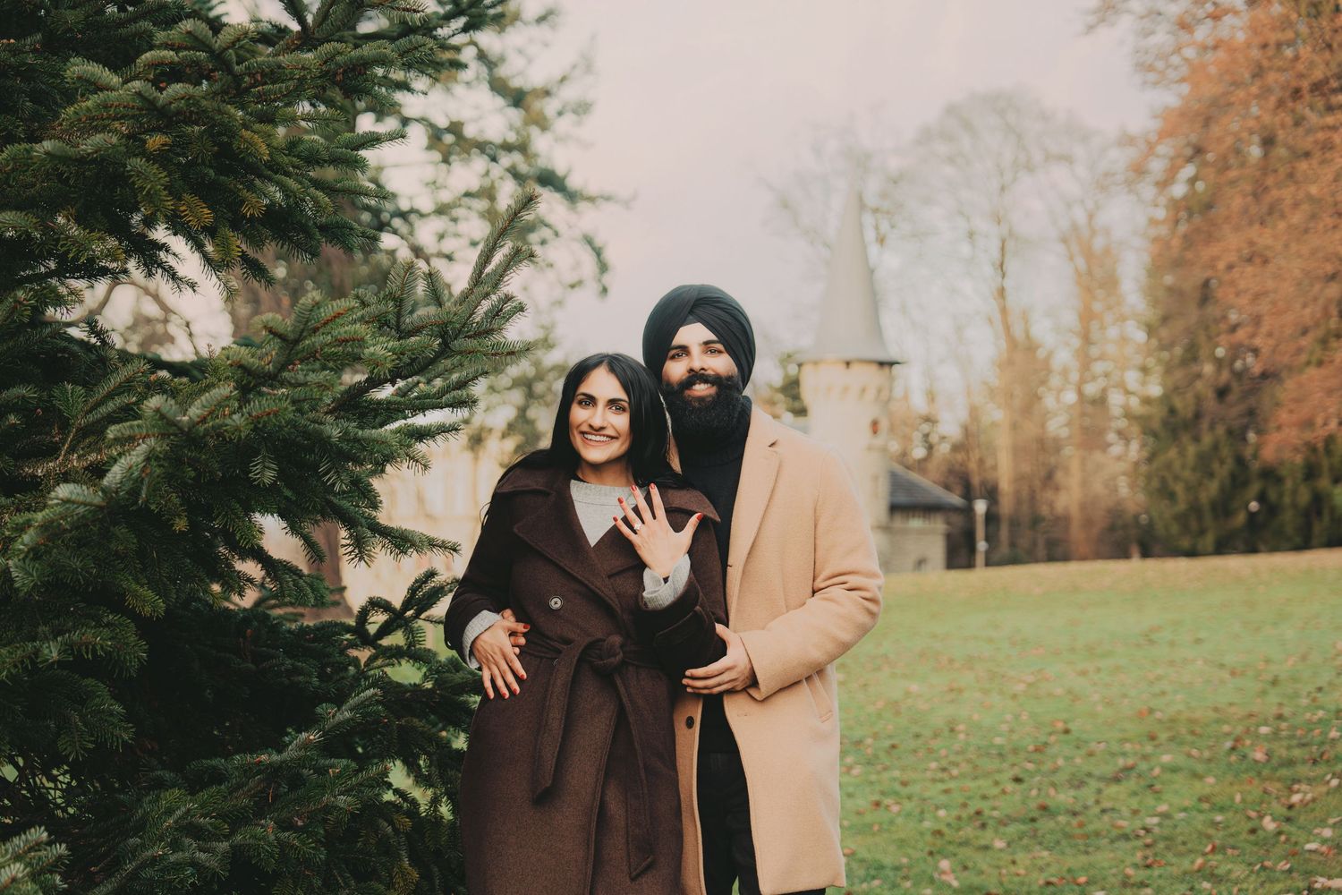 Engaged couple posing by evergreen trees in an autumn park, woman showing ring.