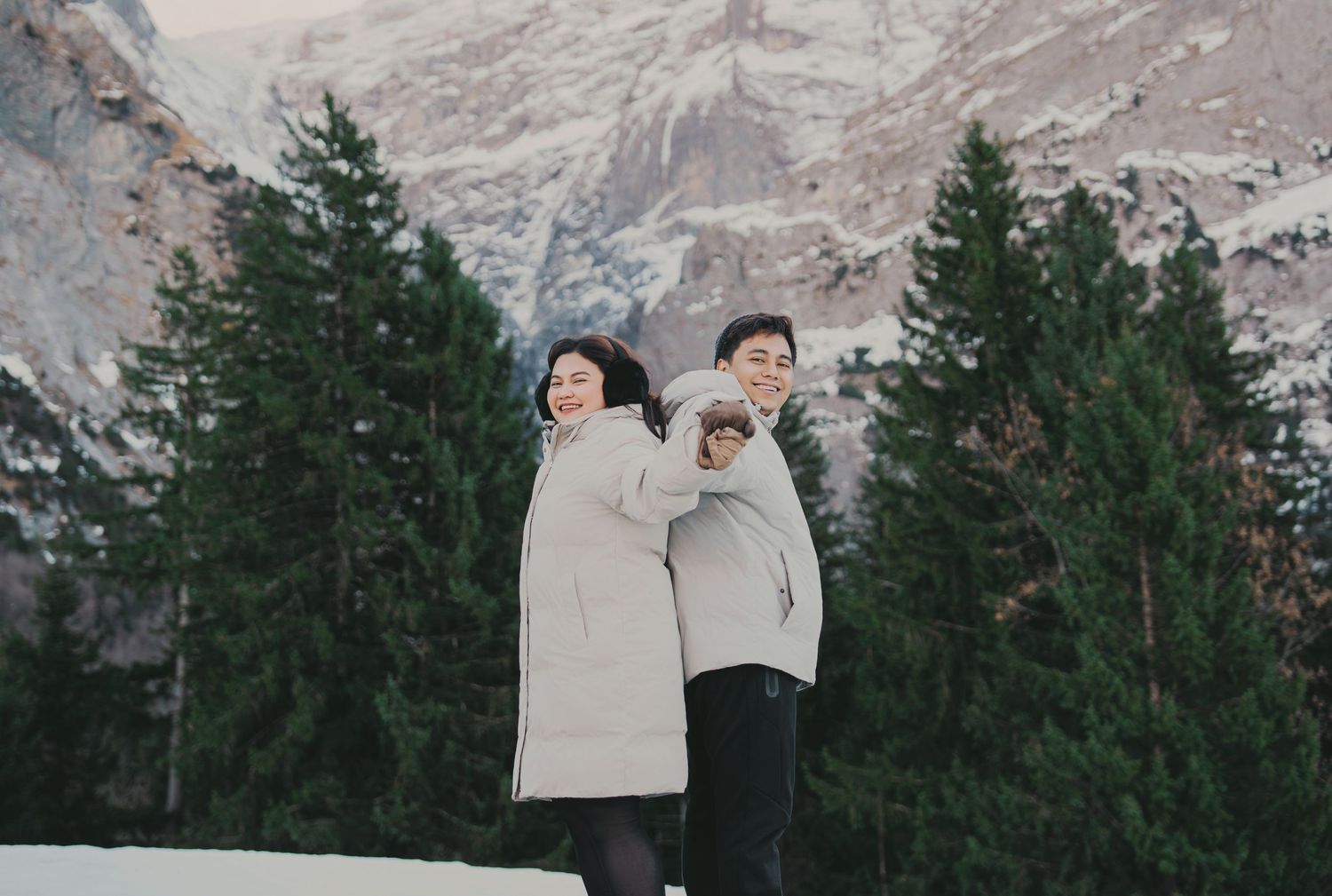 Smiling couple in matching gray coats posing back-to-back with snowy pine trees and mountains behind.