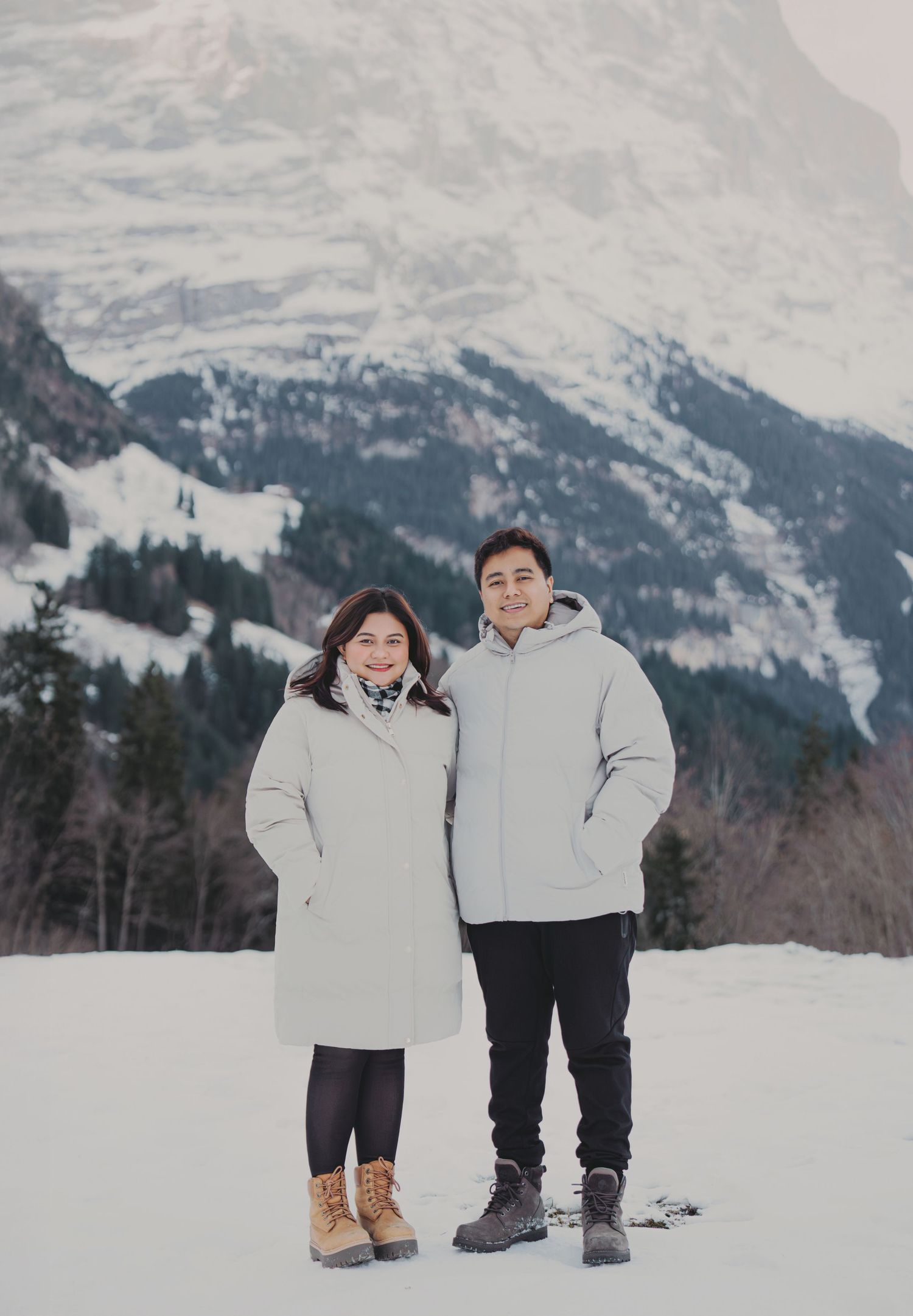 Smiling couple in white winter jackets posing together in front of a dramatic snow-covered alpine mountain.