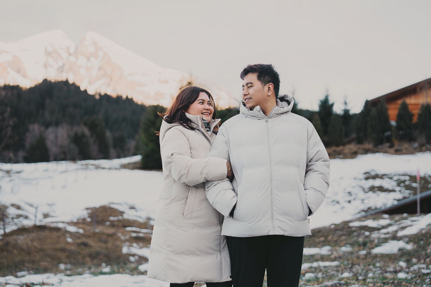 Couple in matching cream puffer jackets smiling at each other in snowy mountain setting at dusk.