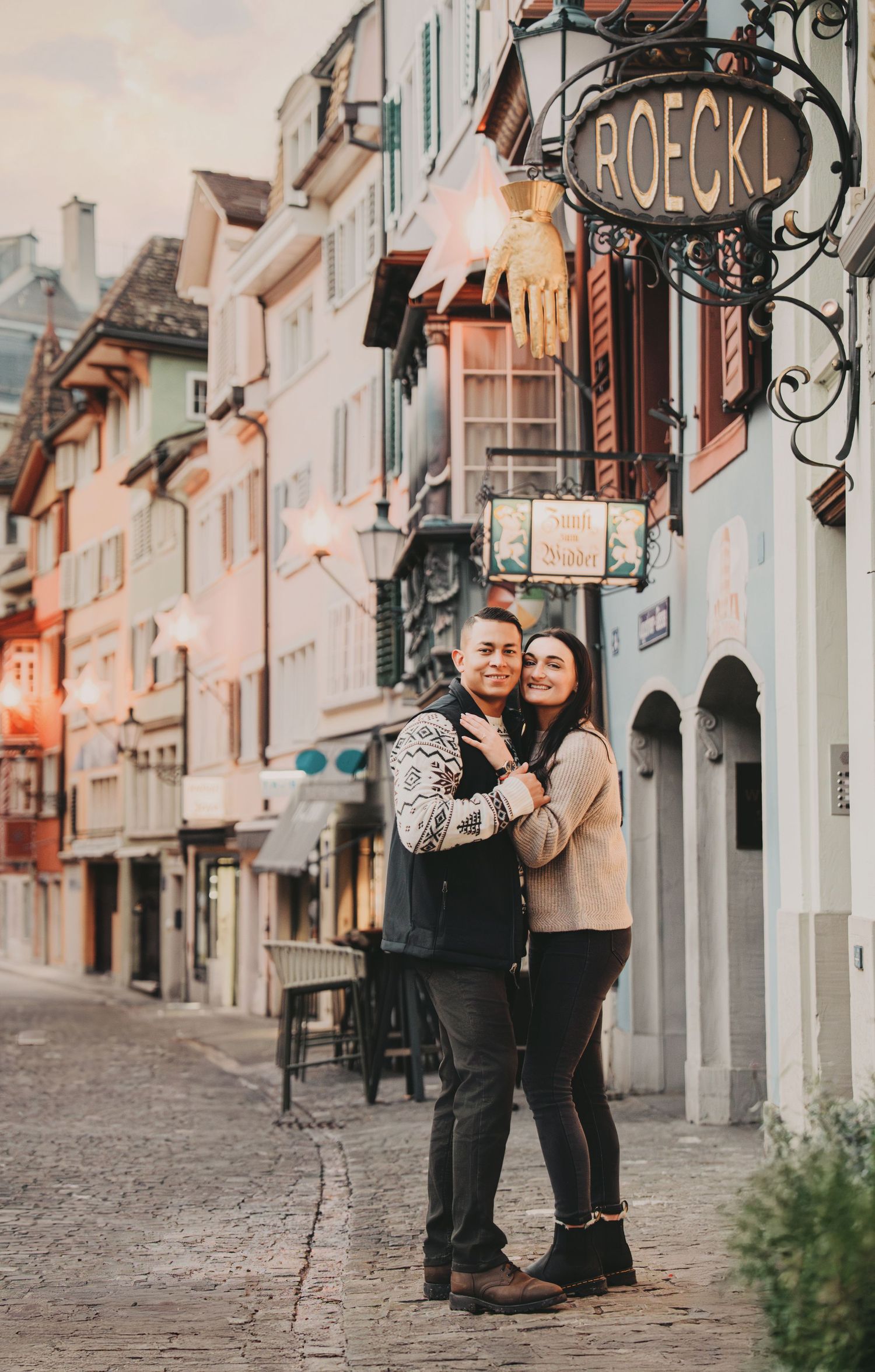 Romantic couple embracing on a charming European cobblestone street near Roeckl shop.