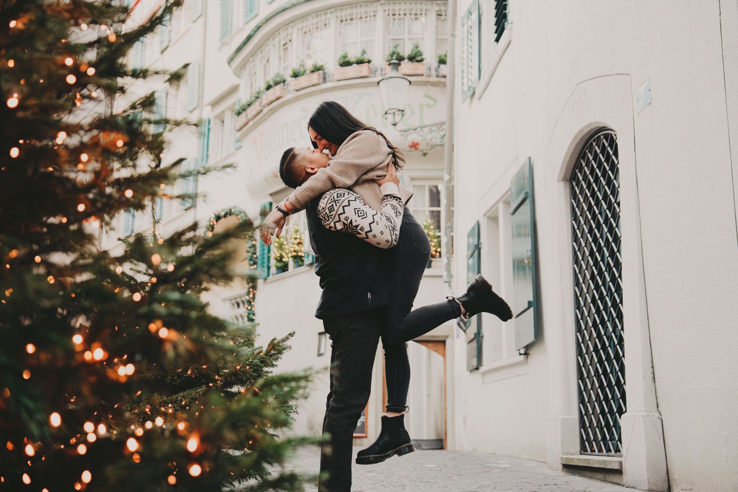 Man lifting woman for a kiss near a decorated Christmas tree on a European street.