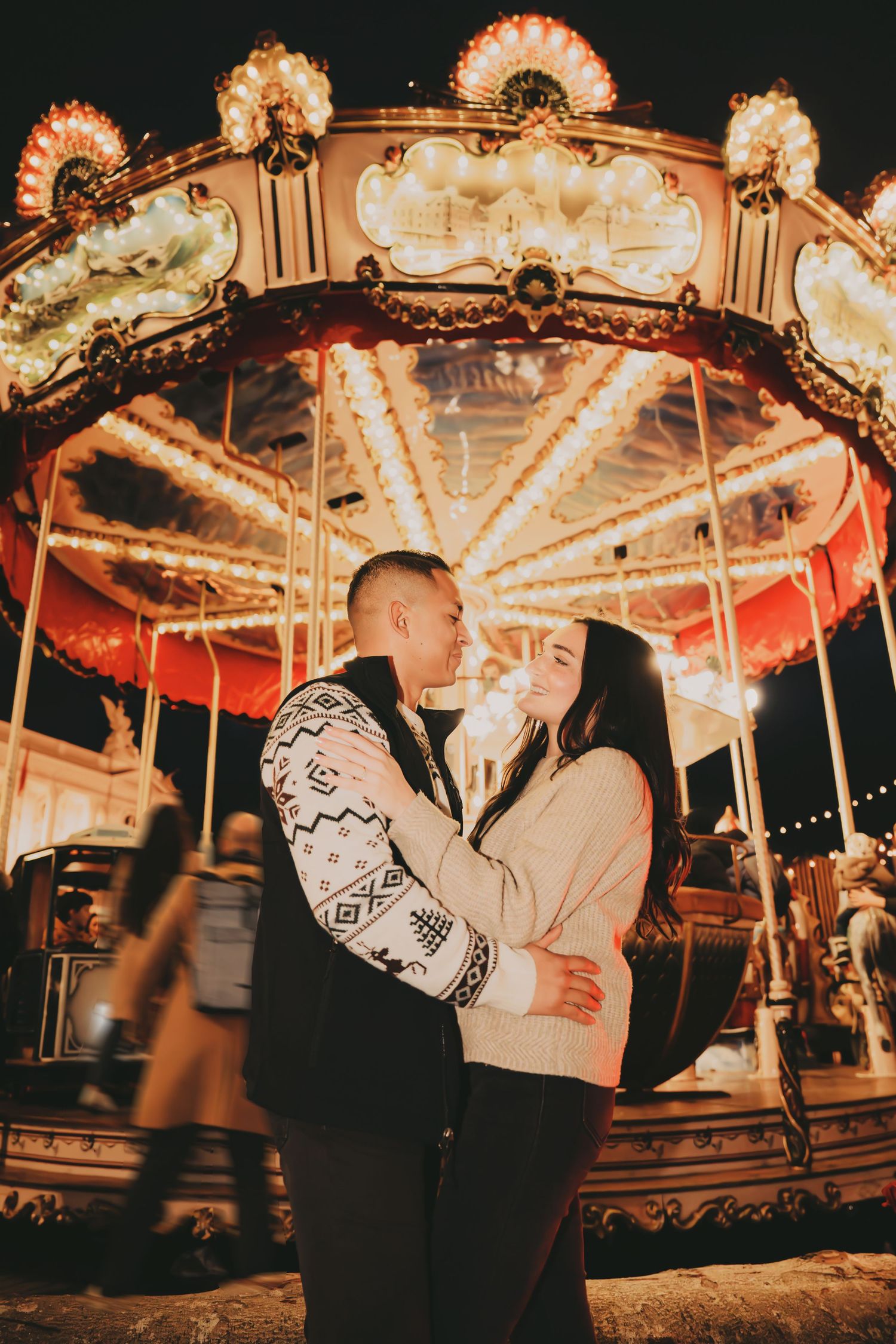 Couple embracing romantically in front of a glowing carousel at night at an amusement park.