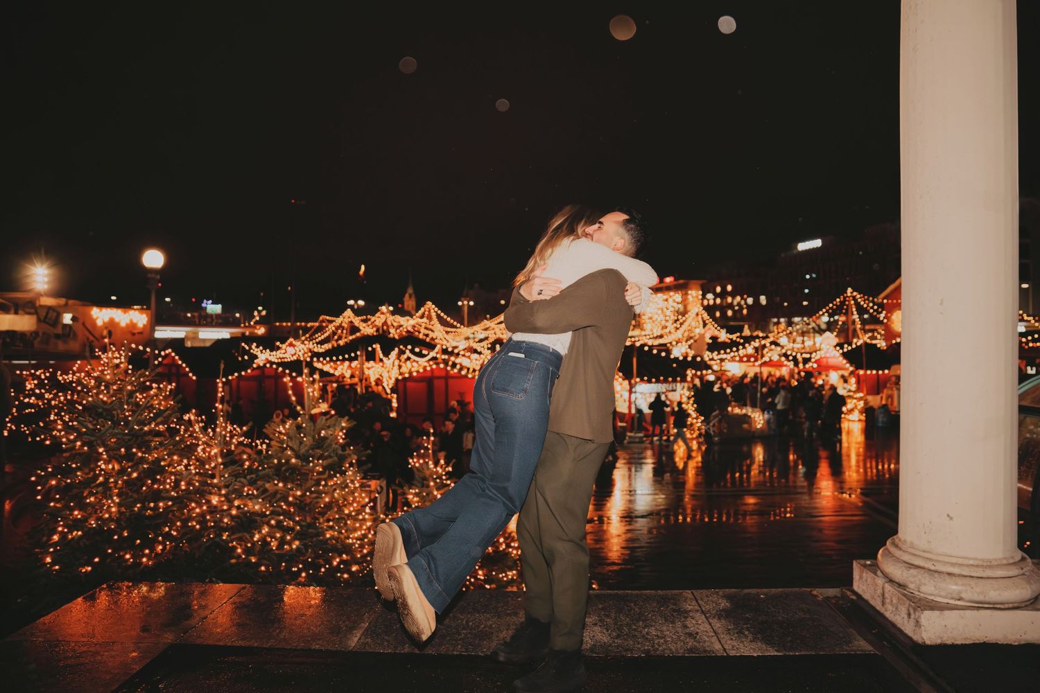 Man lifting woman joyfully at a festive Christmas market with colorful lights and decorations at night.