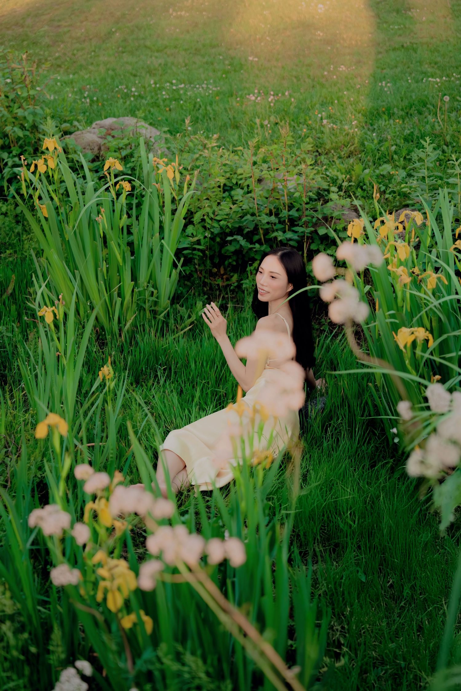 Young woman in yellow dress sitting among yellow irises and wildflowers in lush garden.