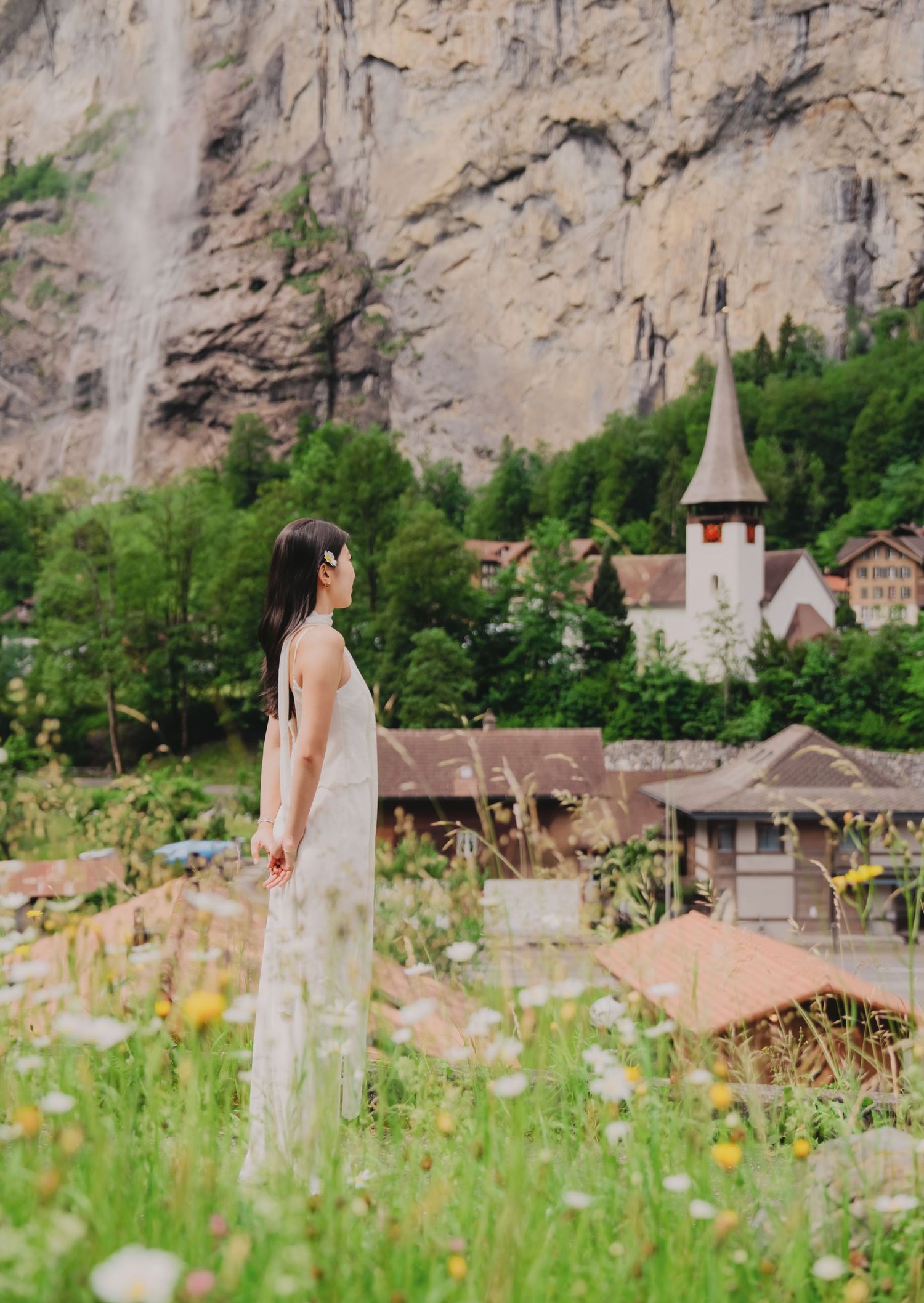 Woman in white dress stands in wildflower meadow with Swiss village church and waterfall backdrop in Lauterbrunnen.